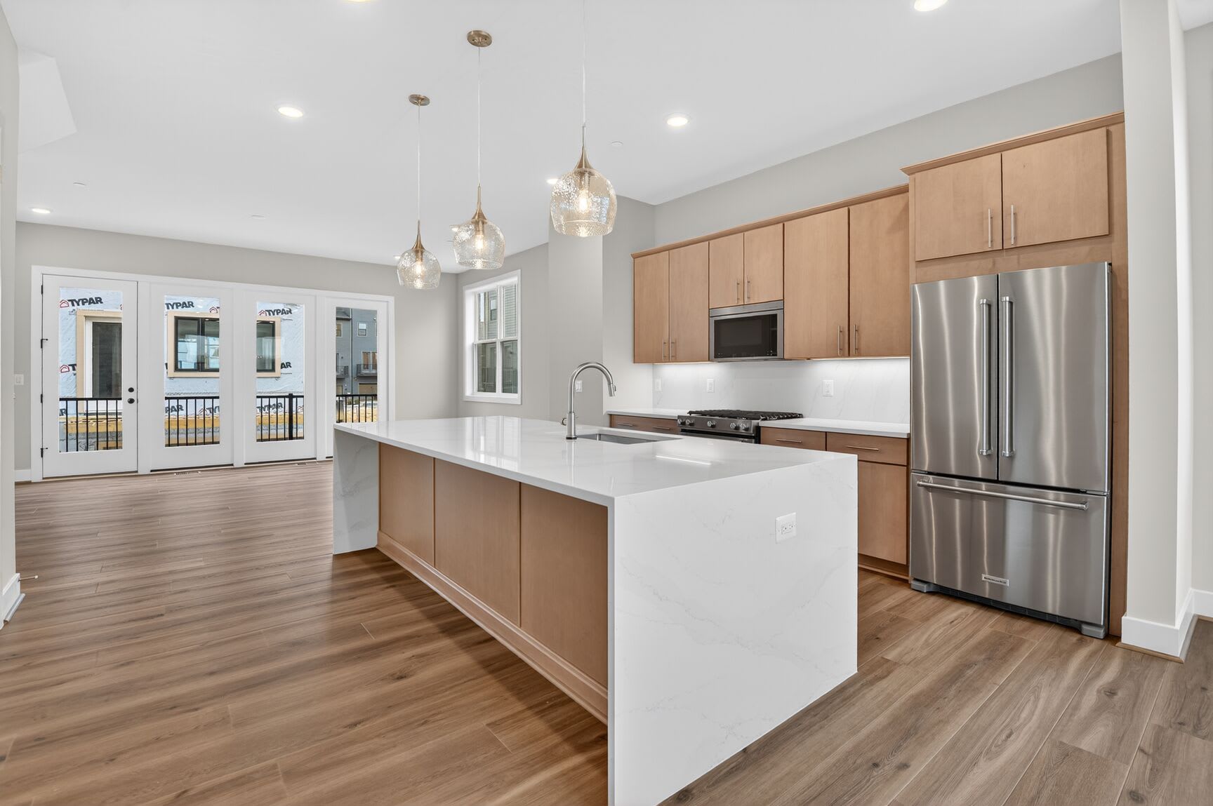 A modern, open-concept kitchen with light wood cabinets, a white countertop, and pendant lighting, leading into a living area with hardwood floors.