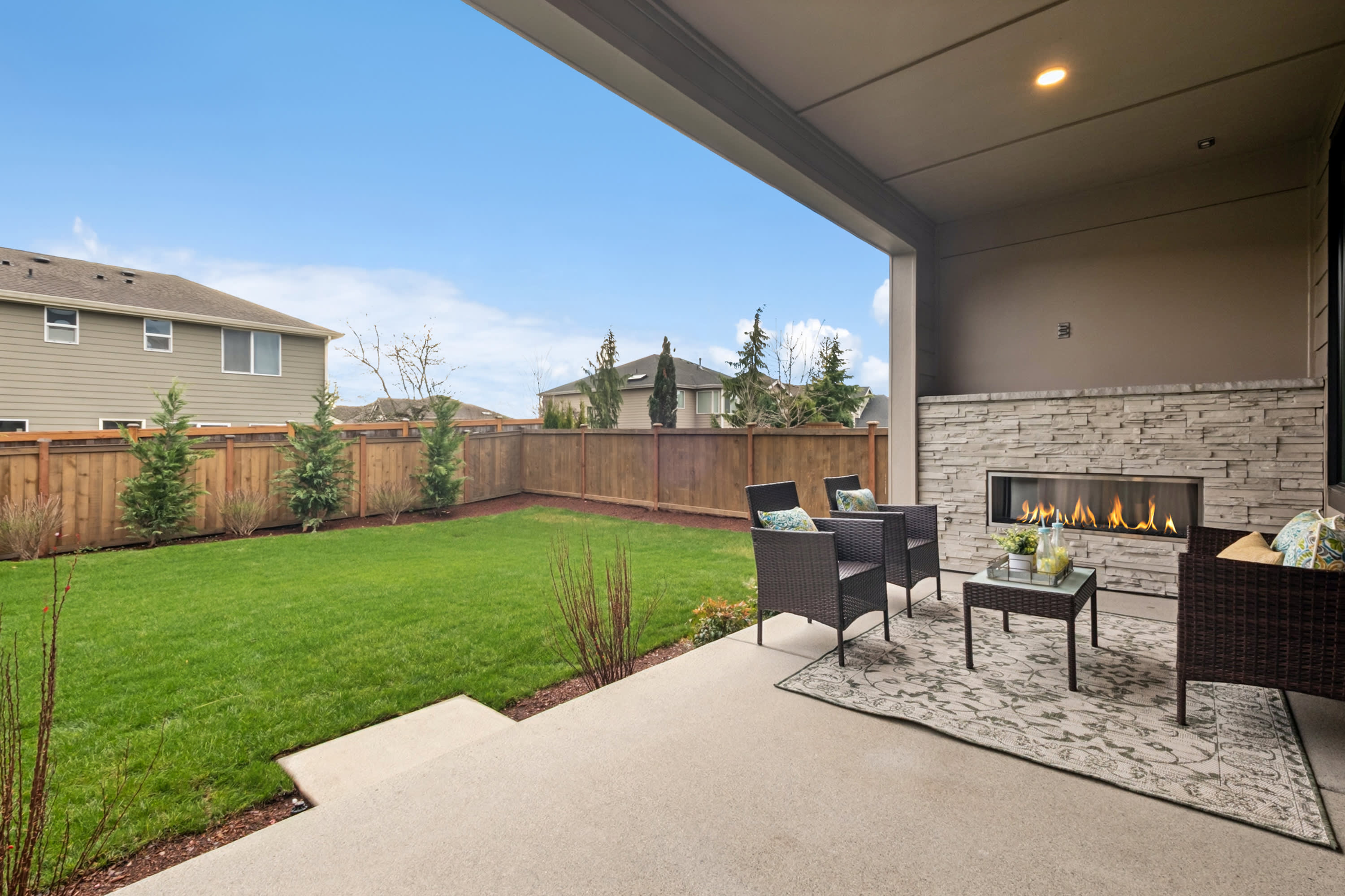 A cozy outdoor living space with a well-manicured lawn, a stone patio, and a fireplace, surrounded by a wooden fence and lush greenery.