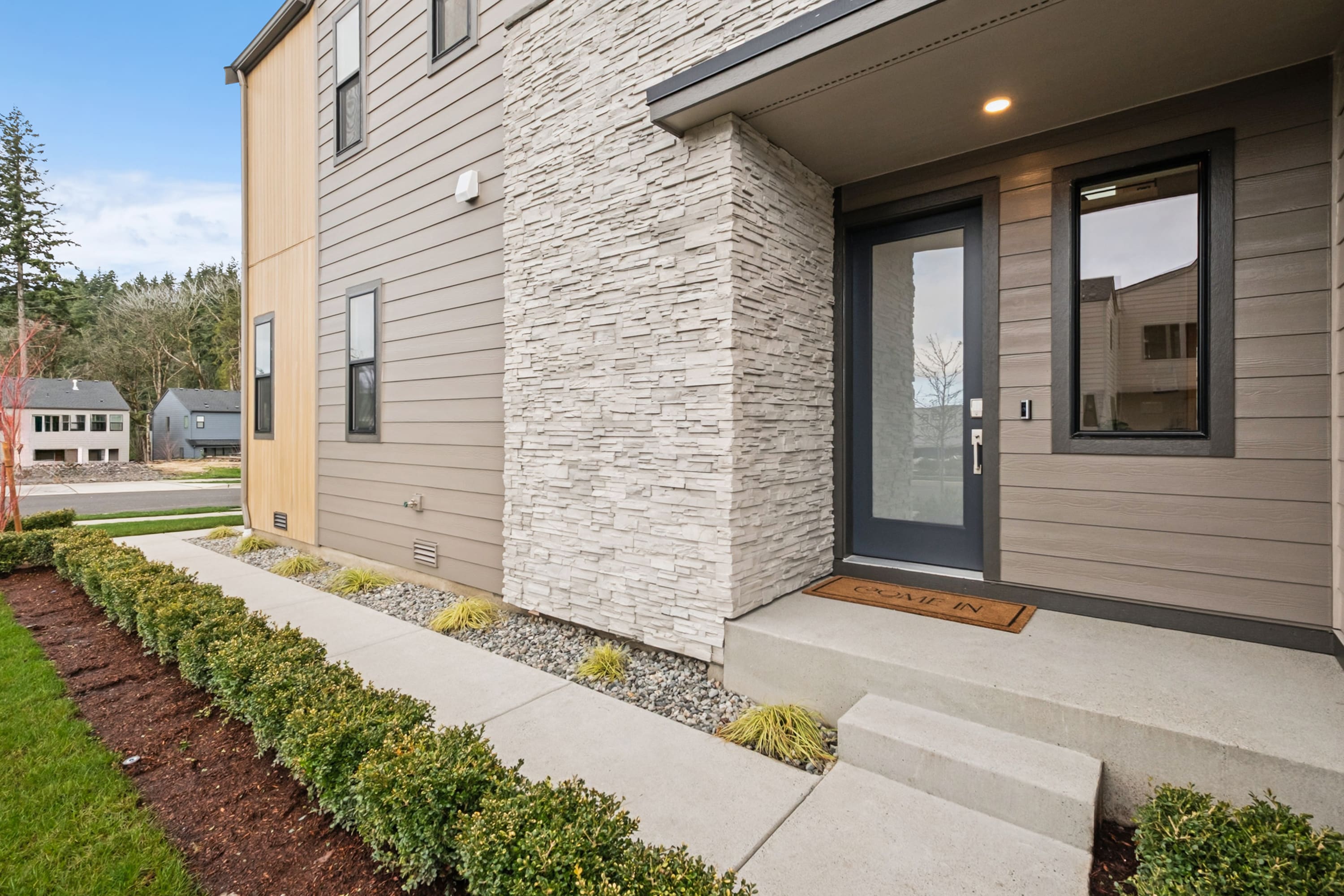 A modern, two-story residential building with a stone facade, a covered entryway, and a well-landscaped front yard featuring shrubs and a paved walkway.