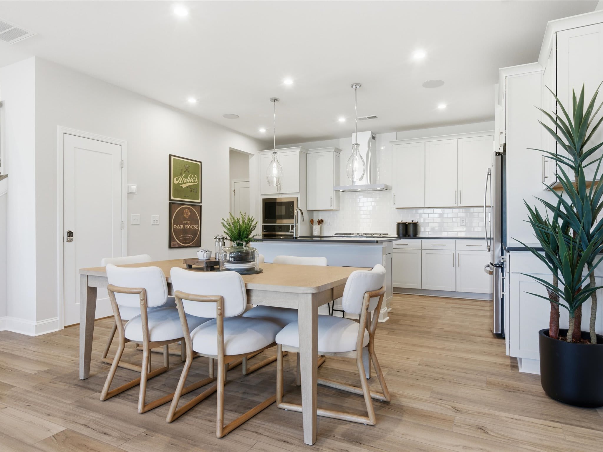 A modern, open-concept kitchen and dining area with white cabinets, wooden furniture, and potted plants, creating a bright and airy atmosphere.