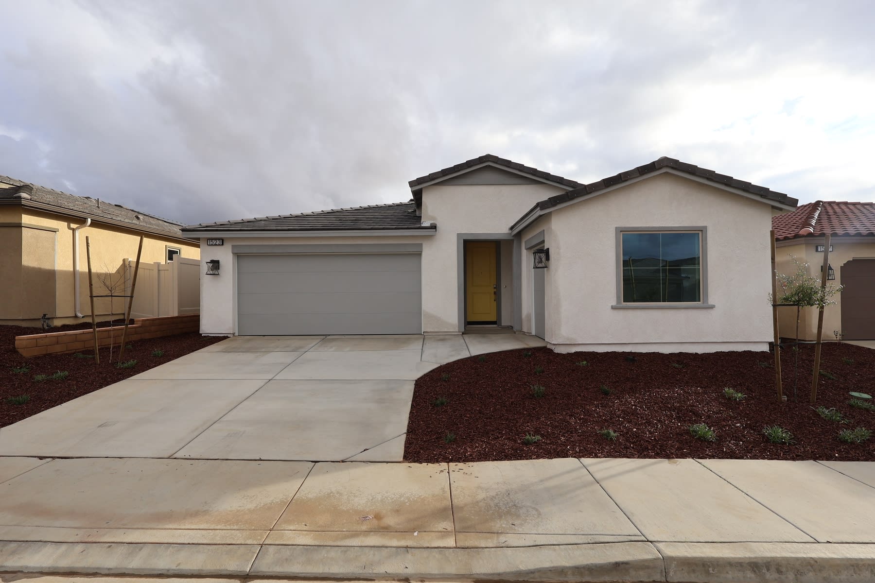 A single-story residential house with a garage, surrounded by a paved driveway and landscaped yard, set against a cloudy sky.