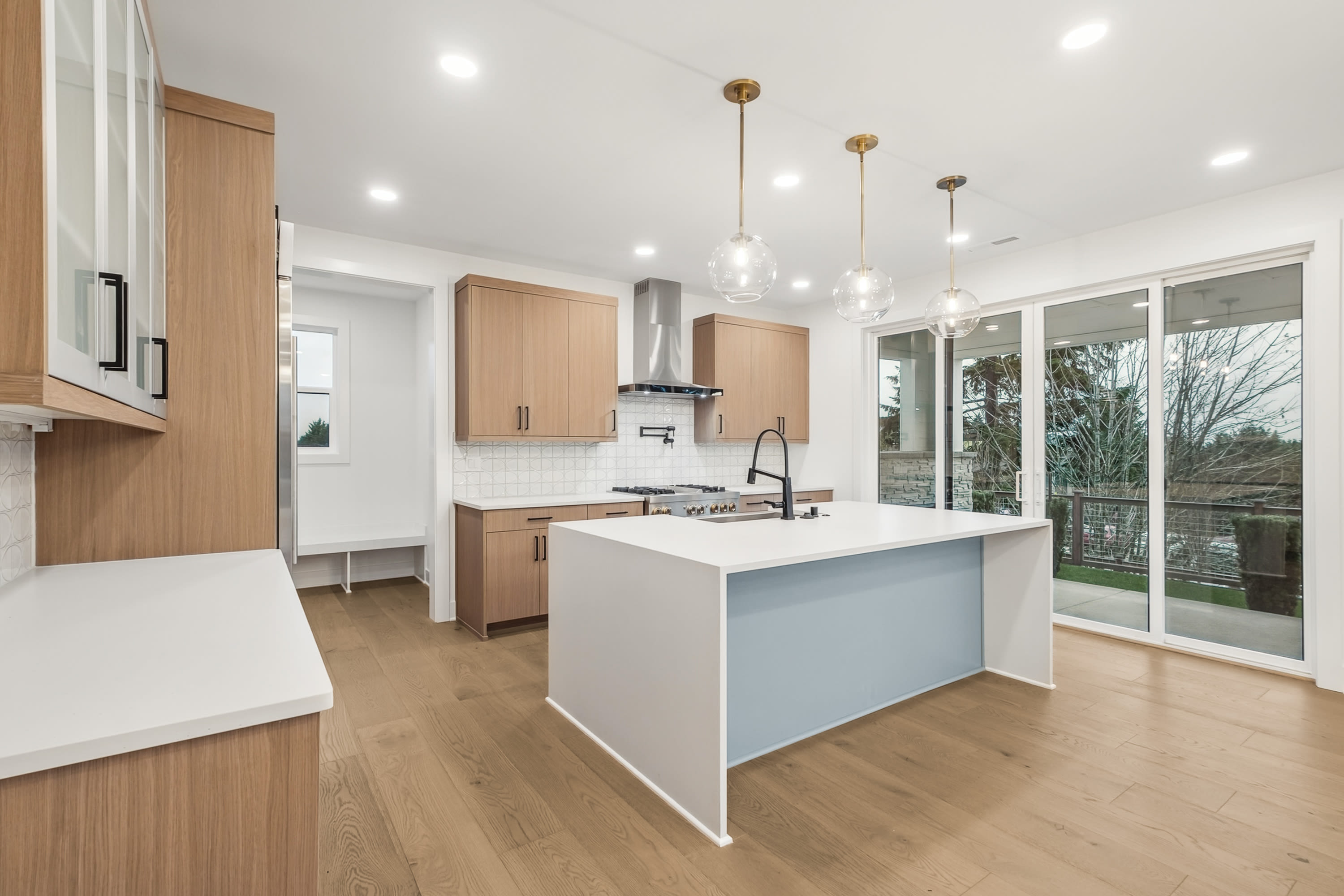 A modern, open-concept kitchen with light wood cabinets, a white island, and pendant lighting fixtures, set against a backdrop of large windows overlooking a wooded outdoor area.