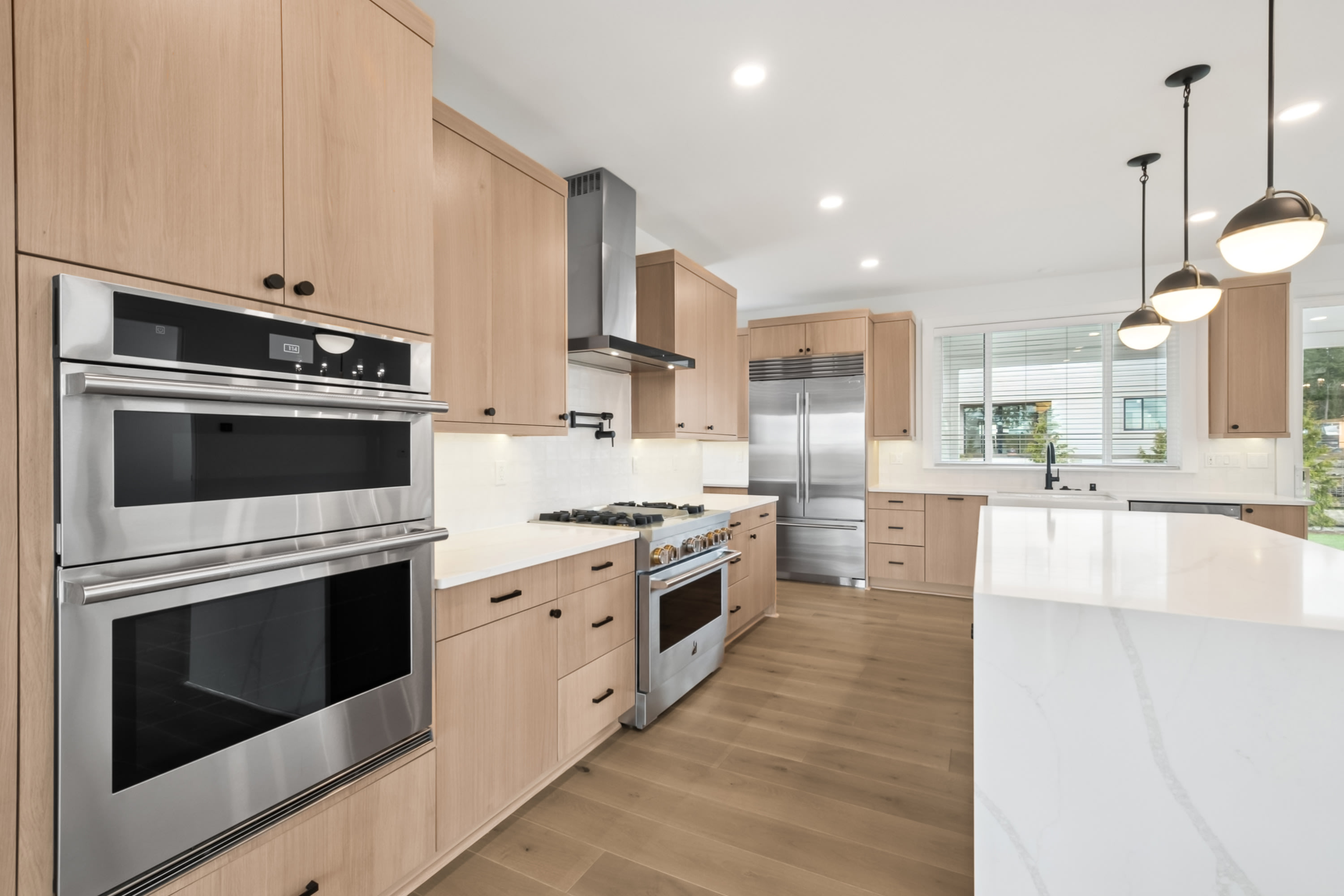 A modern and spacious kitchen with light wood cabinets, stainless steel appliances, and a white countertop that extends into an open-concept living area.