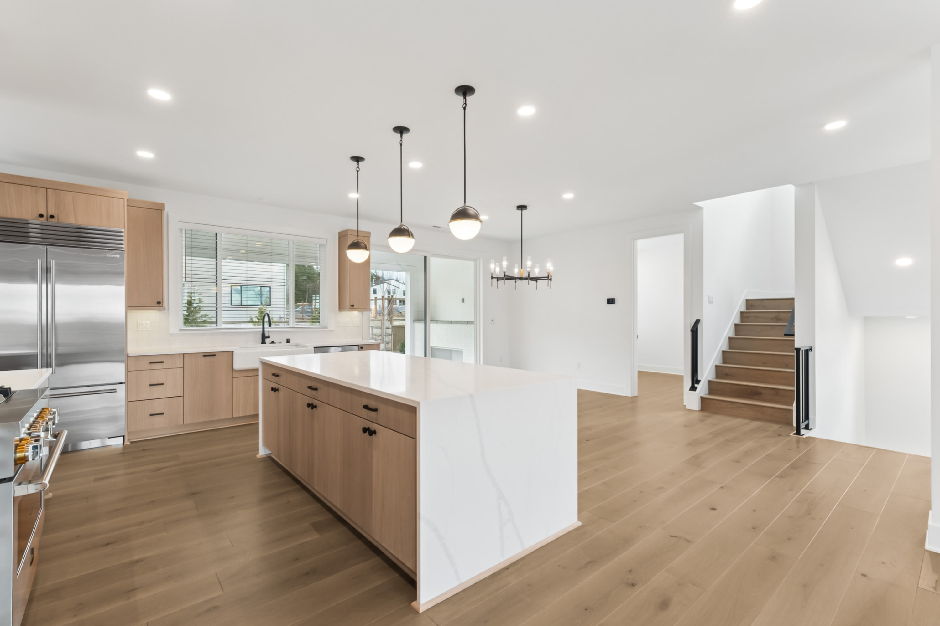 A modern, open-concept kitchen with light wood cabinetry, pendant lighting, and a hardwood floor leading to a staircase in the background.