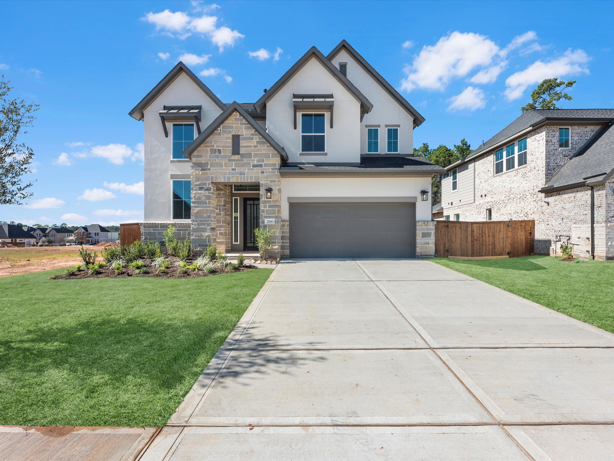 A two-story residential house with a stone and siding exterior, surrounded by a well-manicured lawn and a paved driveway leading to the attached garage, set against a backdrop of a clear blue sky with scattered clouds.