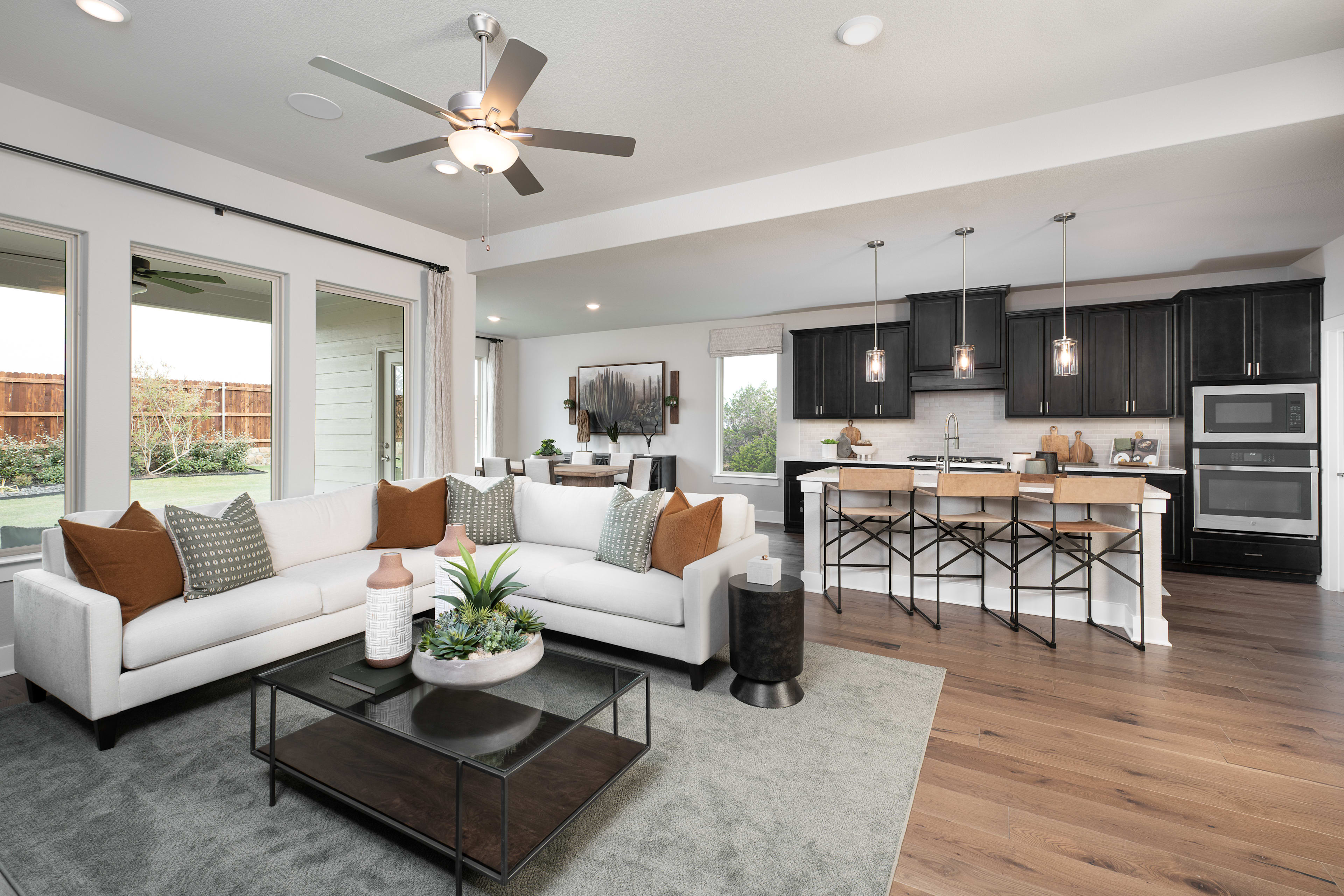 A modern and spacious open-concept living room and kitchen with a white sectional sofa, a wooden coffee table, and a ceiling fan in the background.