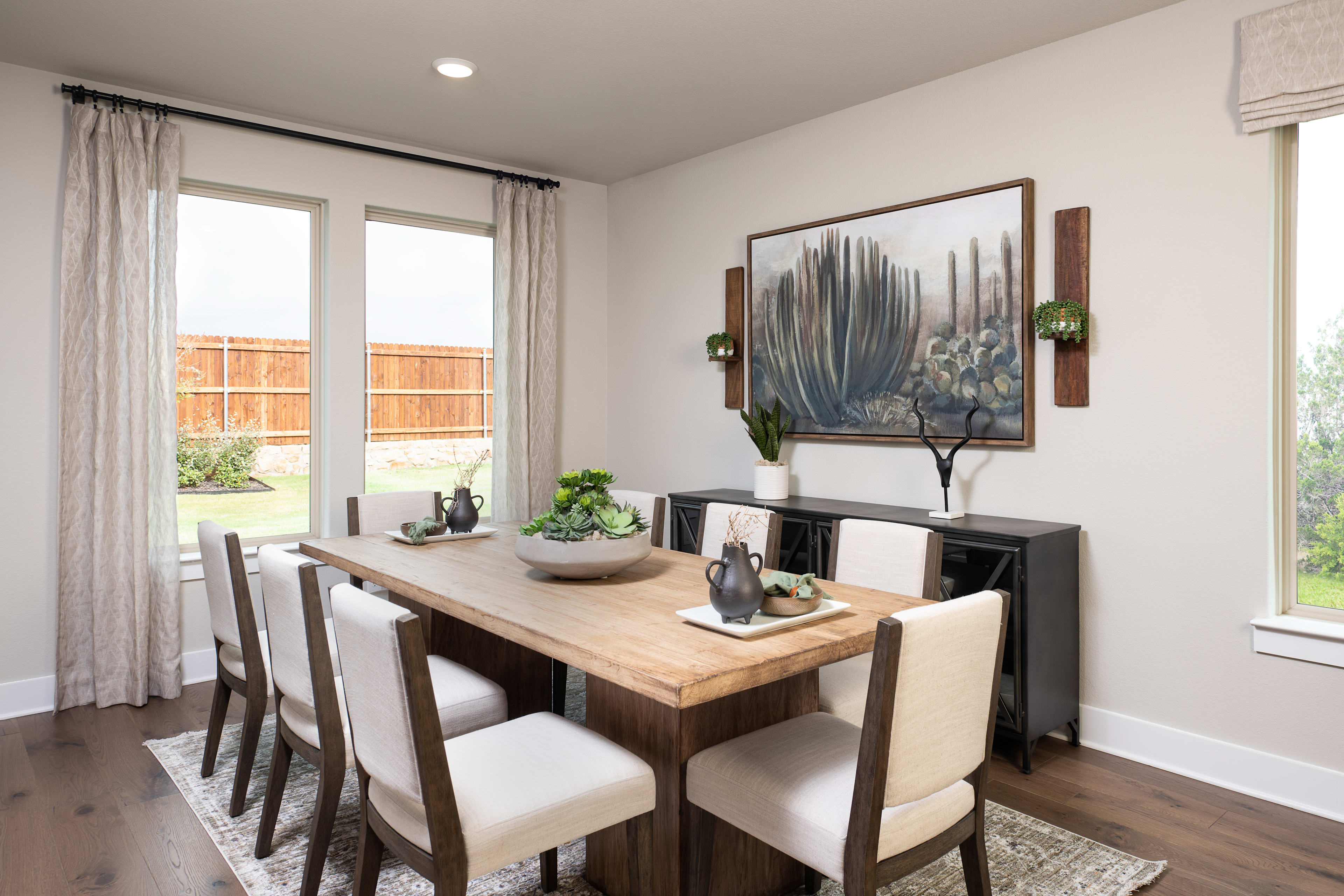 A modern and cozy dining room with a wooden table, upholstered chairs, and a large abstract painting on the wall, complemented by potted plants and a wooden sideboard.