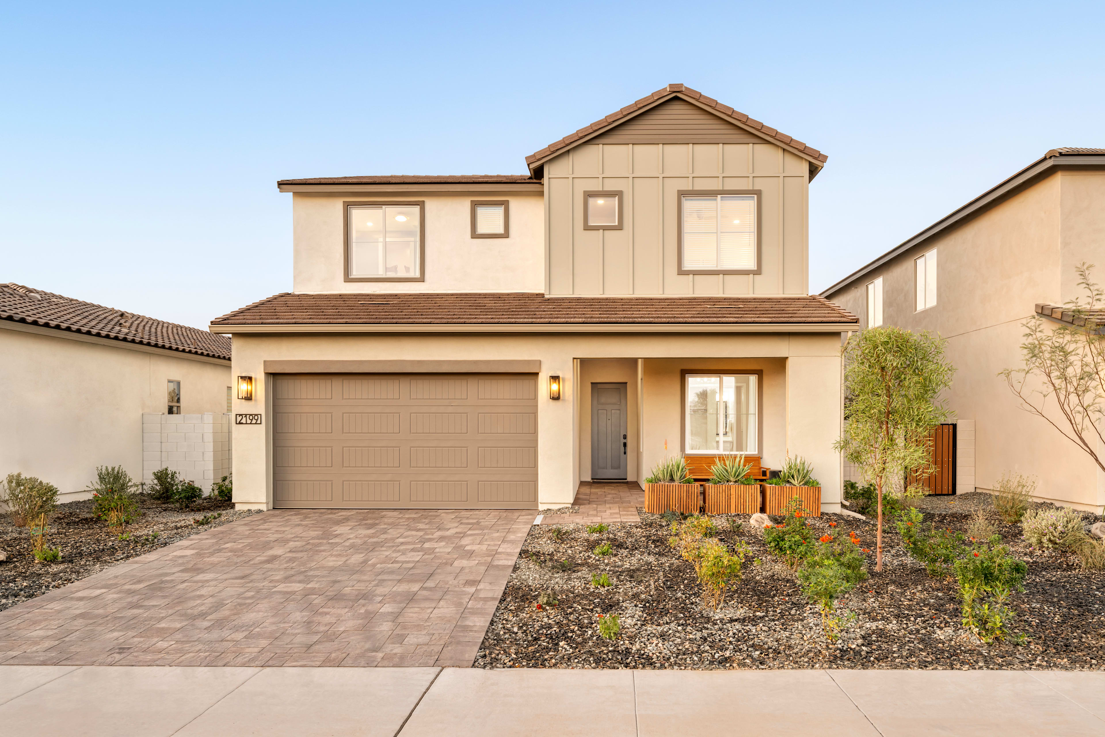 A two-story beige house with a garage, surrounded by a landscaped yard with plants and flowers in the foreground.