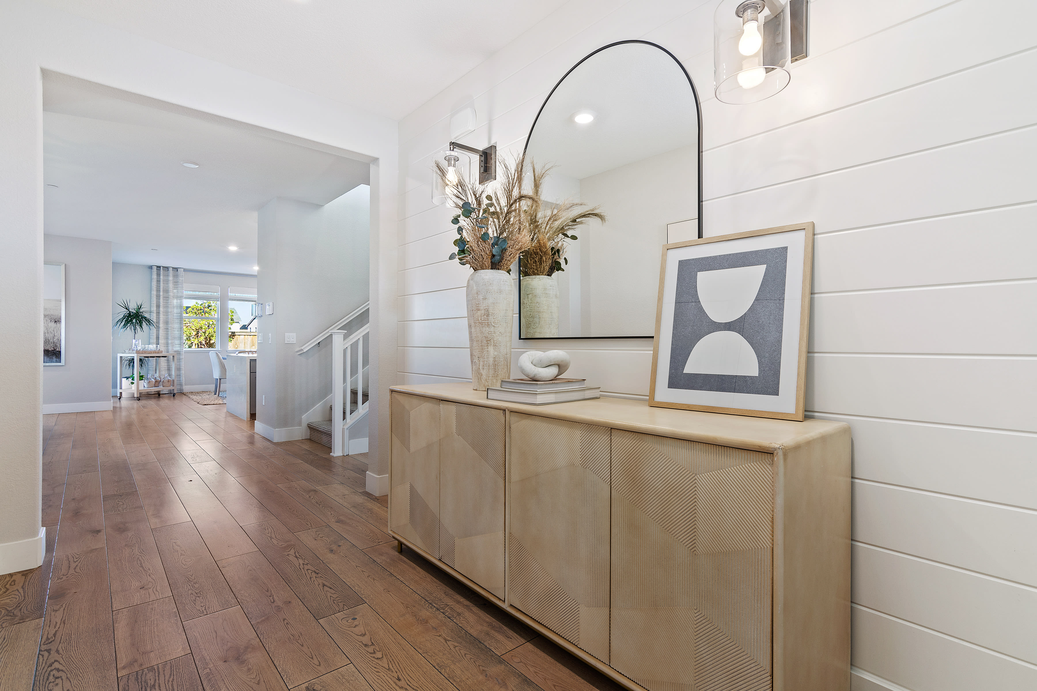 A spacious and well-lit hallway with a wooden console table, a large arched mirror, and a framed abstract artwork on the wall, leading to an open room in the background.