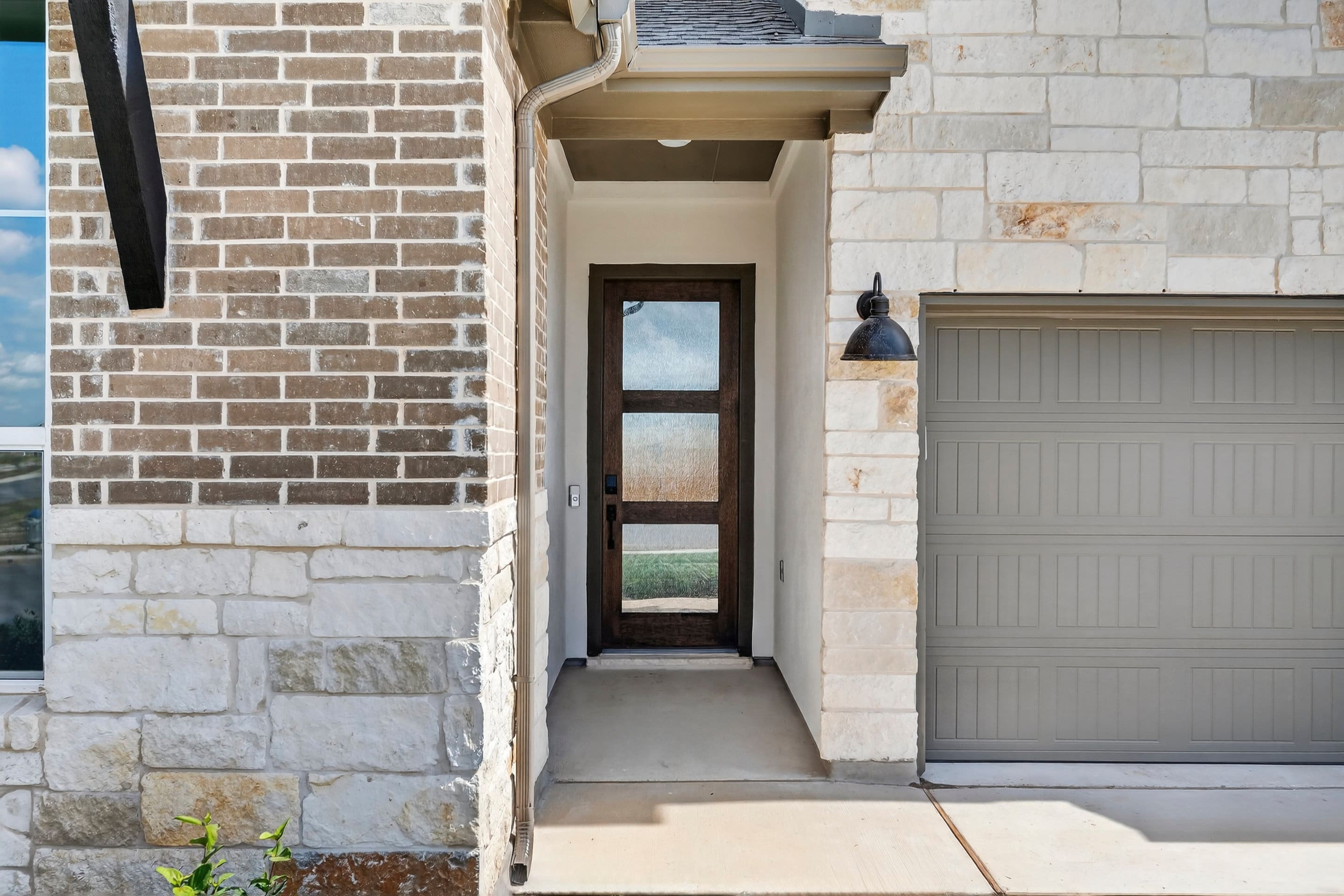 A brick and stone exterior with a wooden door and garage door, leading to a paved walkway and a grassy area in the background.