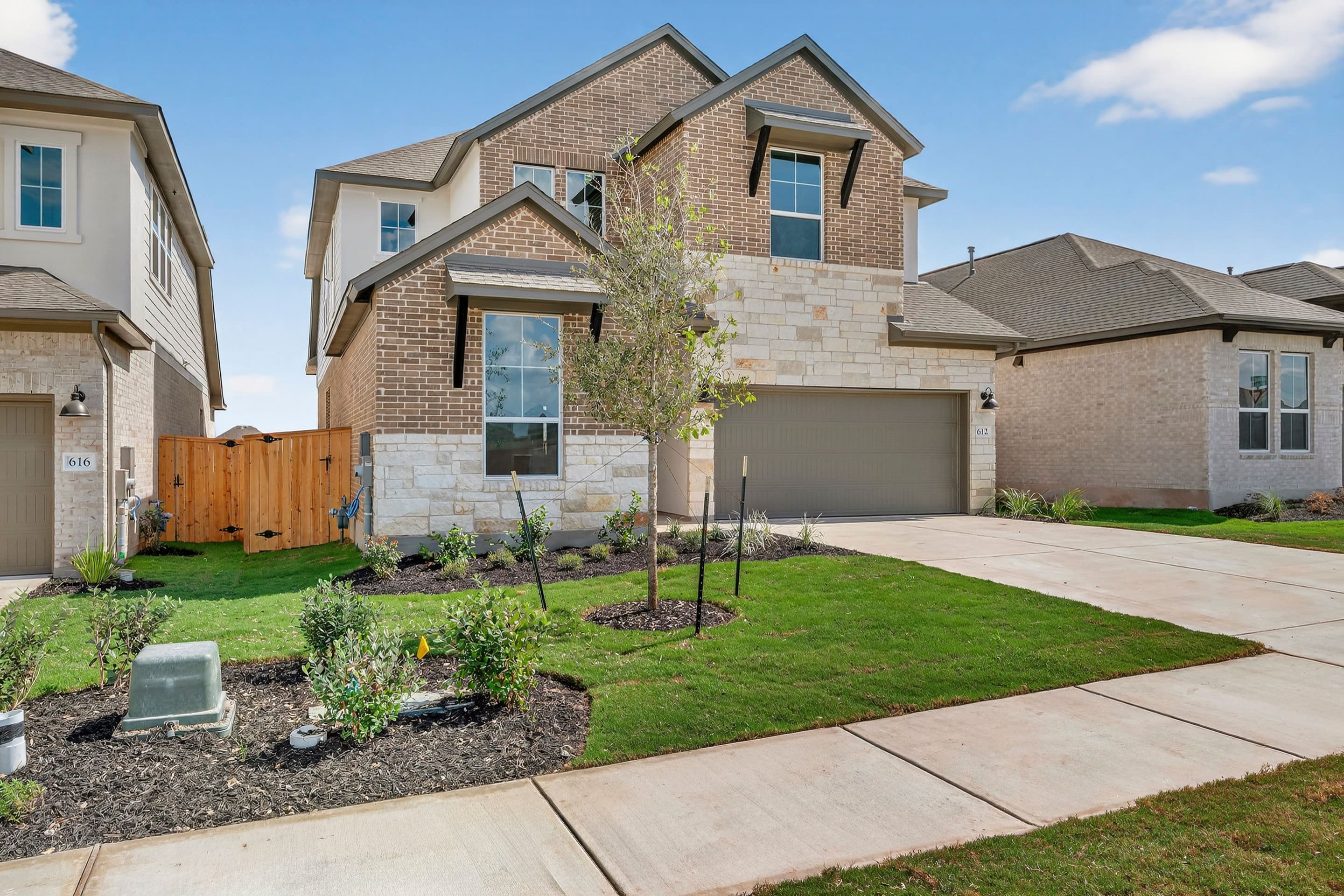 A two-story brick house with a well-manicured lawn, surrounded by other residential buildings, set against a clear blue sky with some clouds.