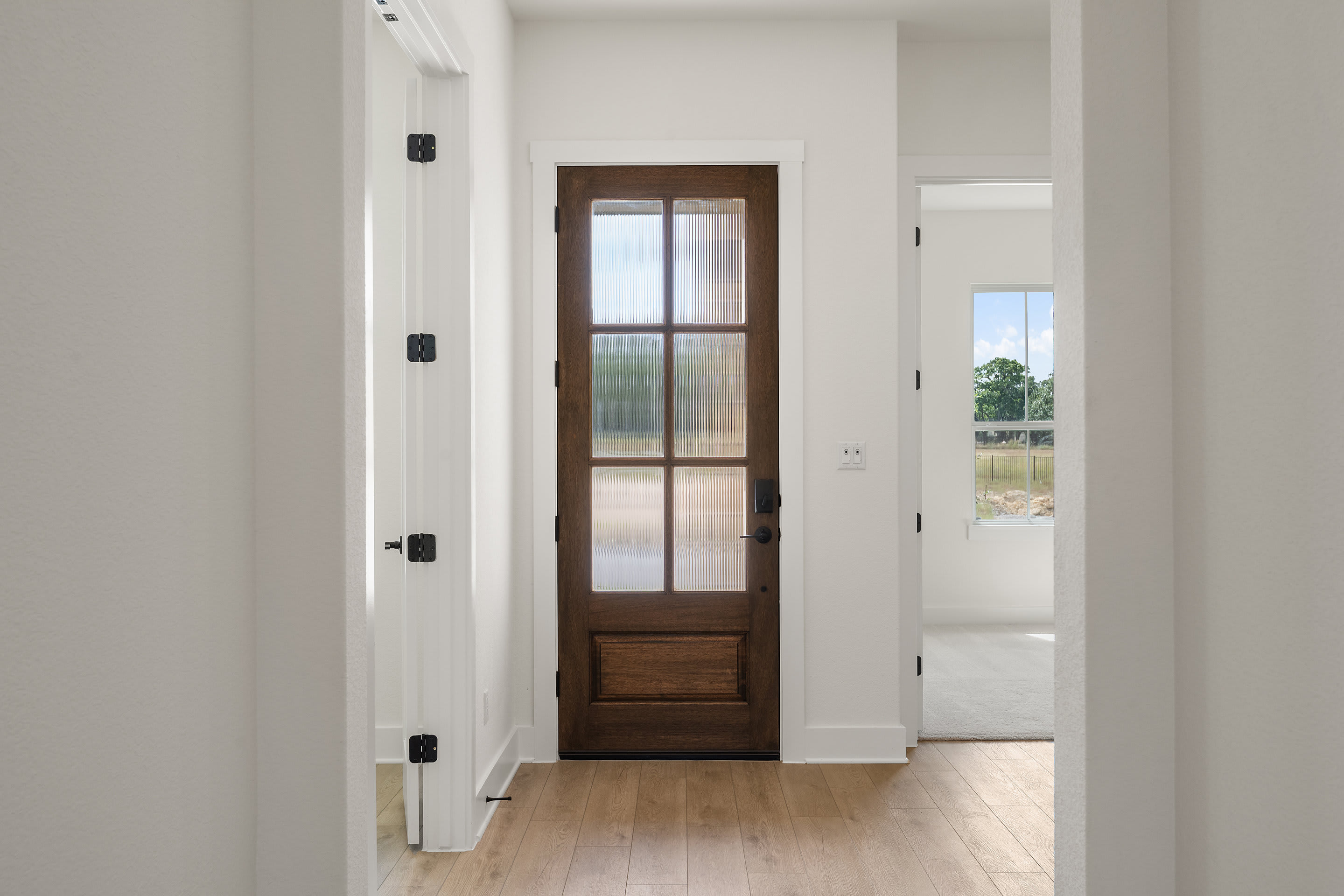 A hallway with a wooden door featuring a glass panel, leading to a bright and open space beyond.