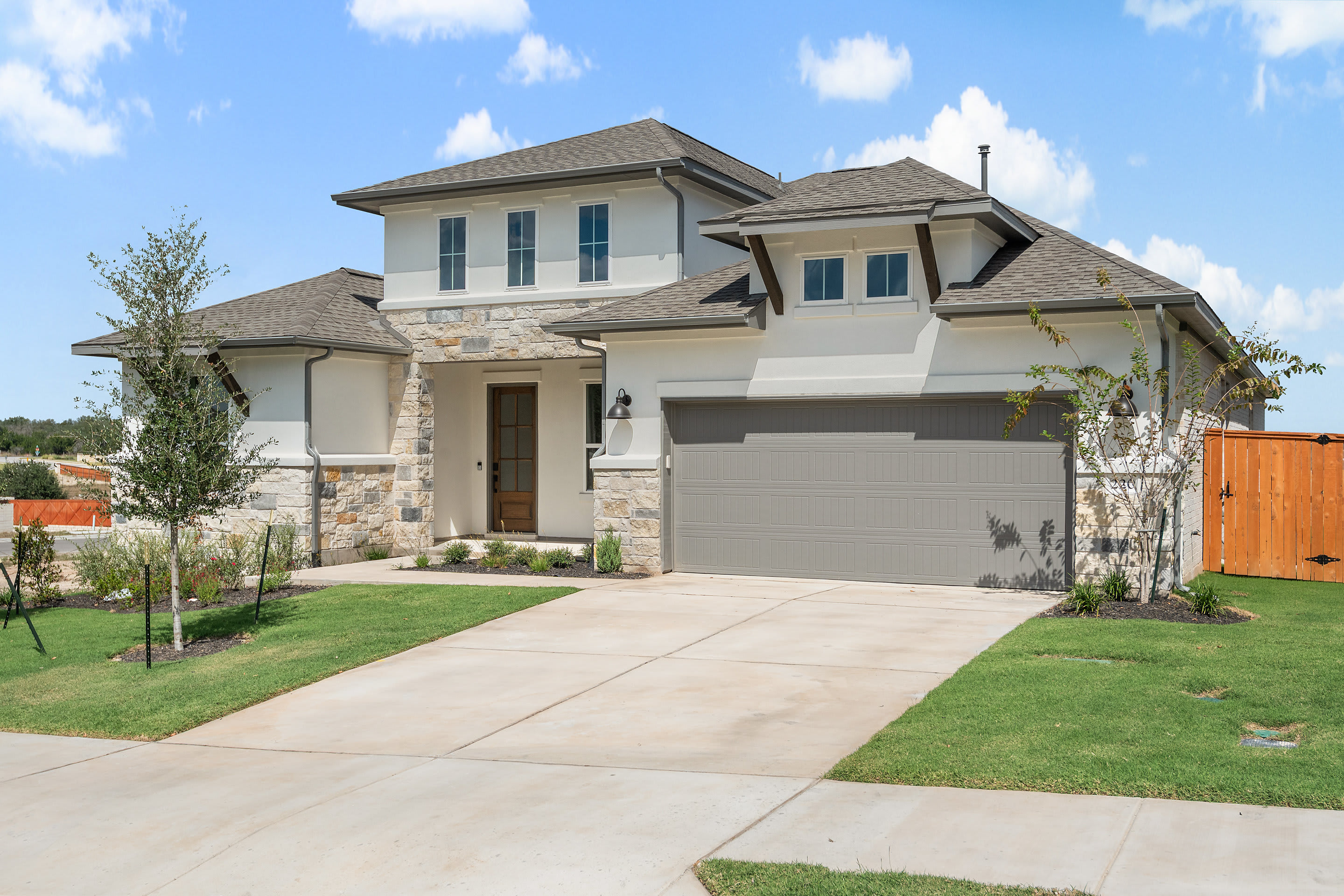 A two-story residential house with a stone exterior, a garage, and a well-manicured lawn in the foreground, set against a blue sky with scattered clouds.