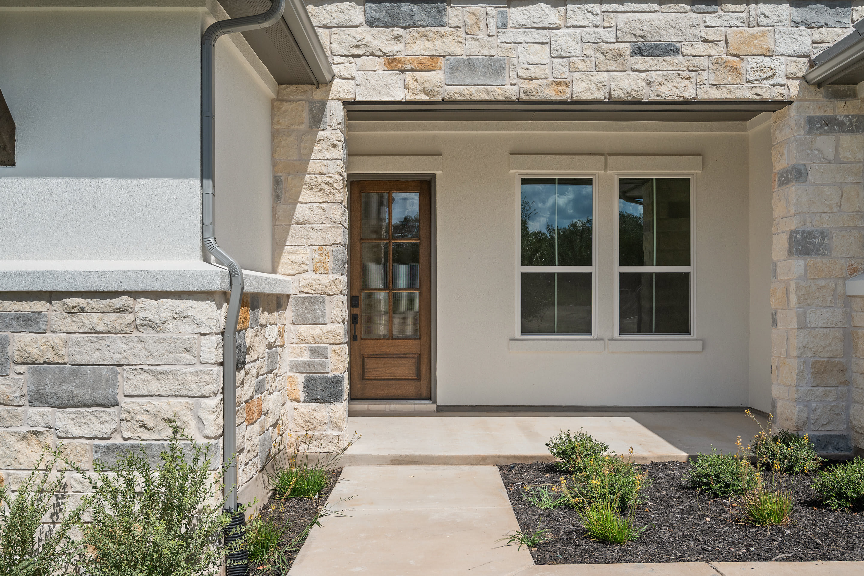 A stone-clad entryway with a wooden door and windows, surrounded by landscaped greenery, leading to a modern residential structure.