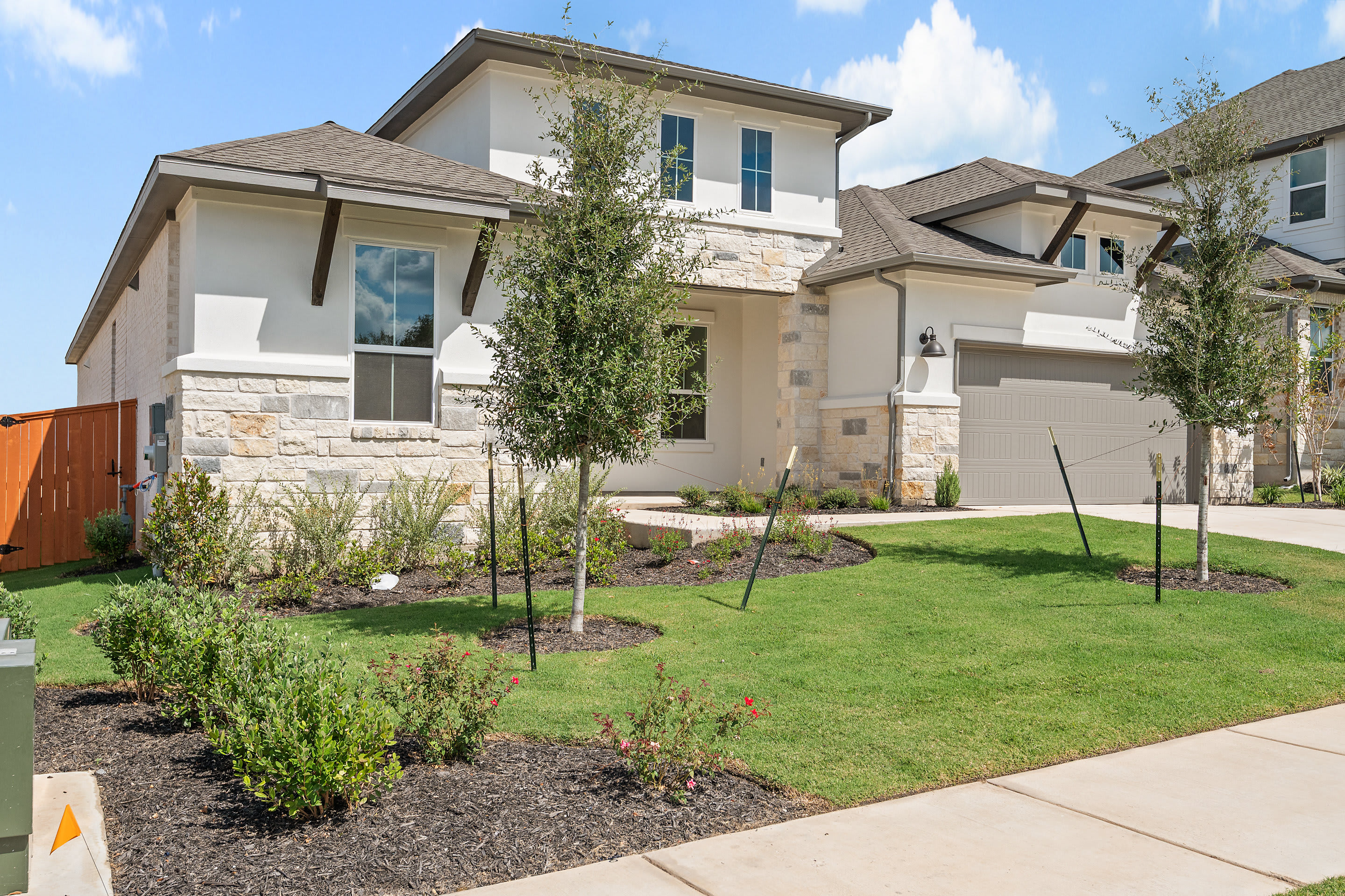 A well-manicured residential neighborhood with a two-story house featuring a stone exterior, a lush green lawn, and various landscaping elements such as trees and shrubs.