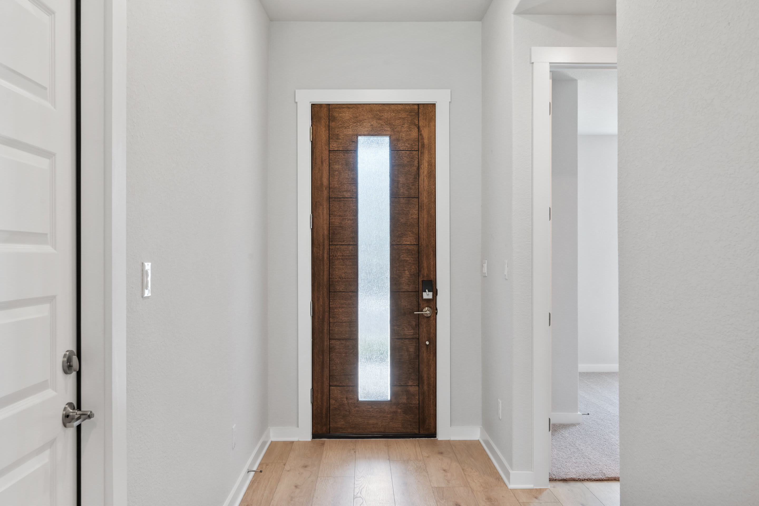 A narrow wooden door with a vertical window panel stands in a bright, white-walled hallway, leading to another room beyond.