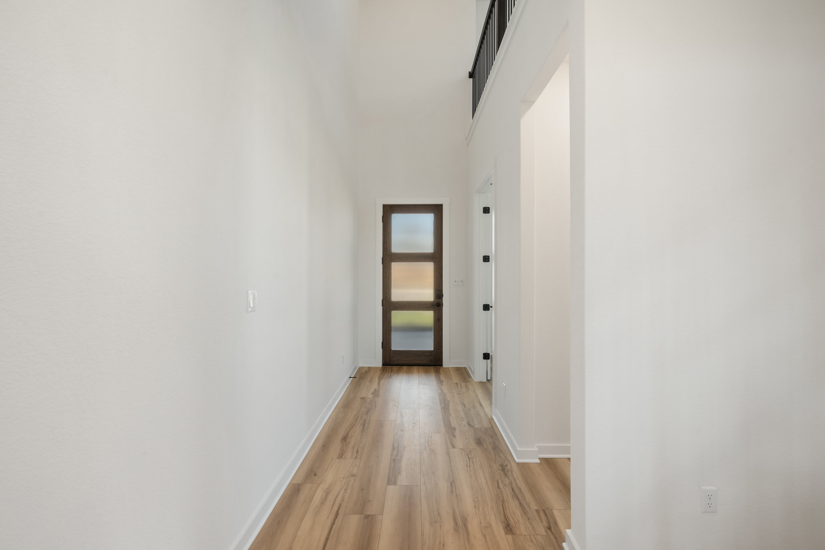 A long, narrow hallway with white walls and a wooden floor, featuring a small window with colored glass panes.