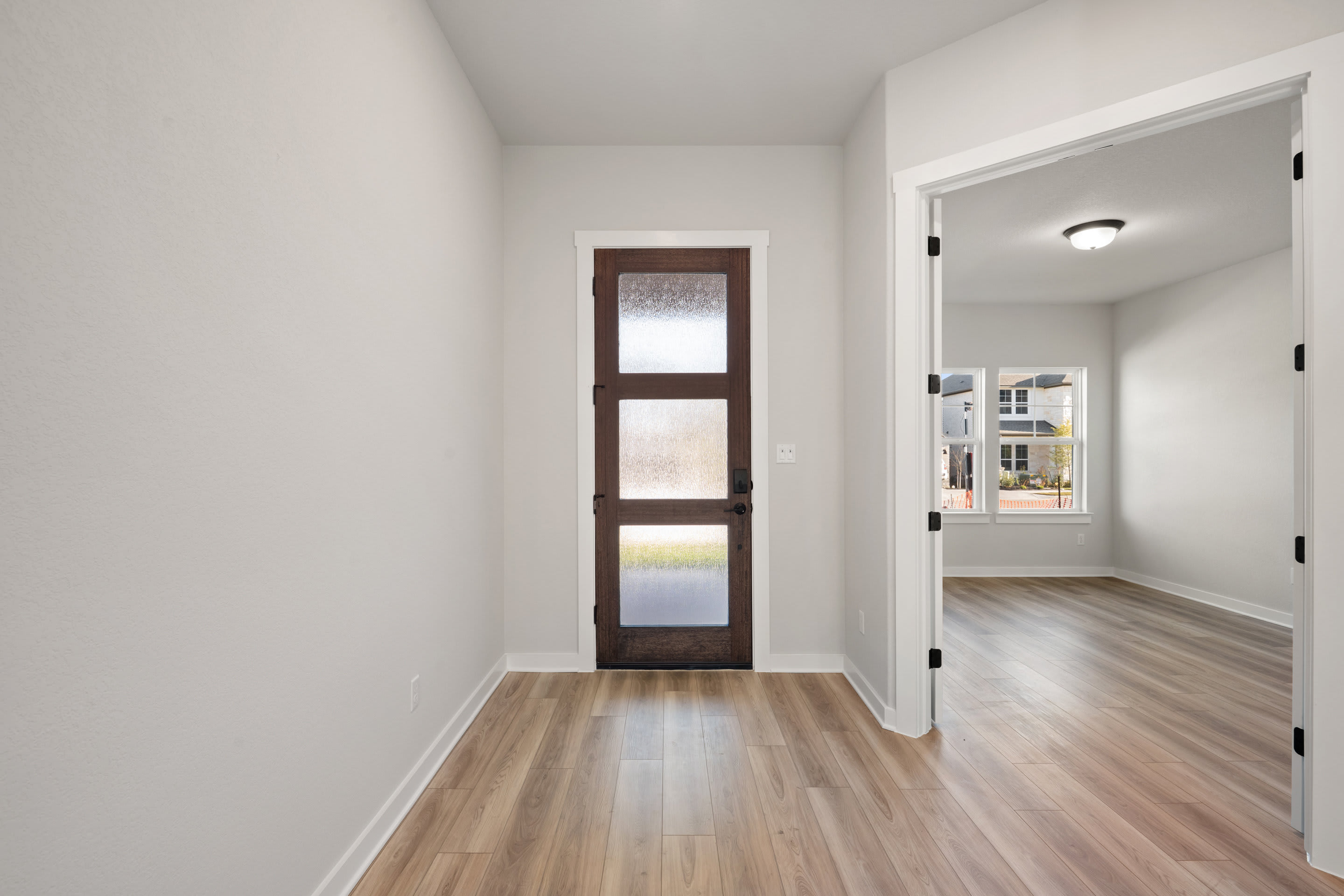 A spacious and well-lit hallway with a wooden door and a hardwood floor, leading to another room visible in the background.