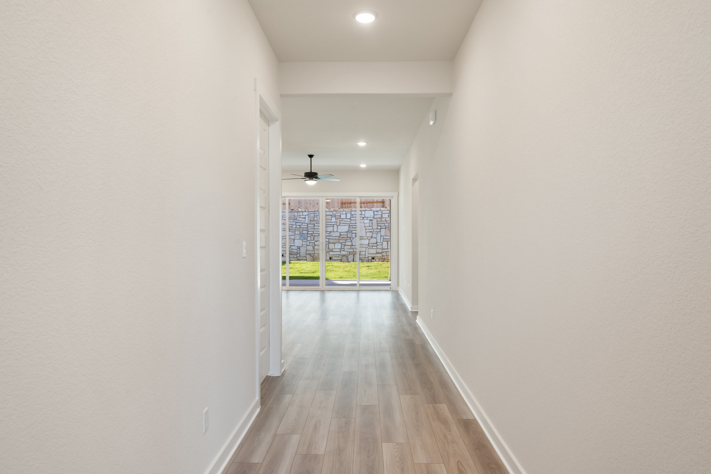 A long, well-lit hallway with white walls and a wooden floor, leading to a window at the end that provides natural light and a view of the outdoors.