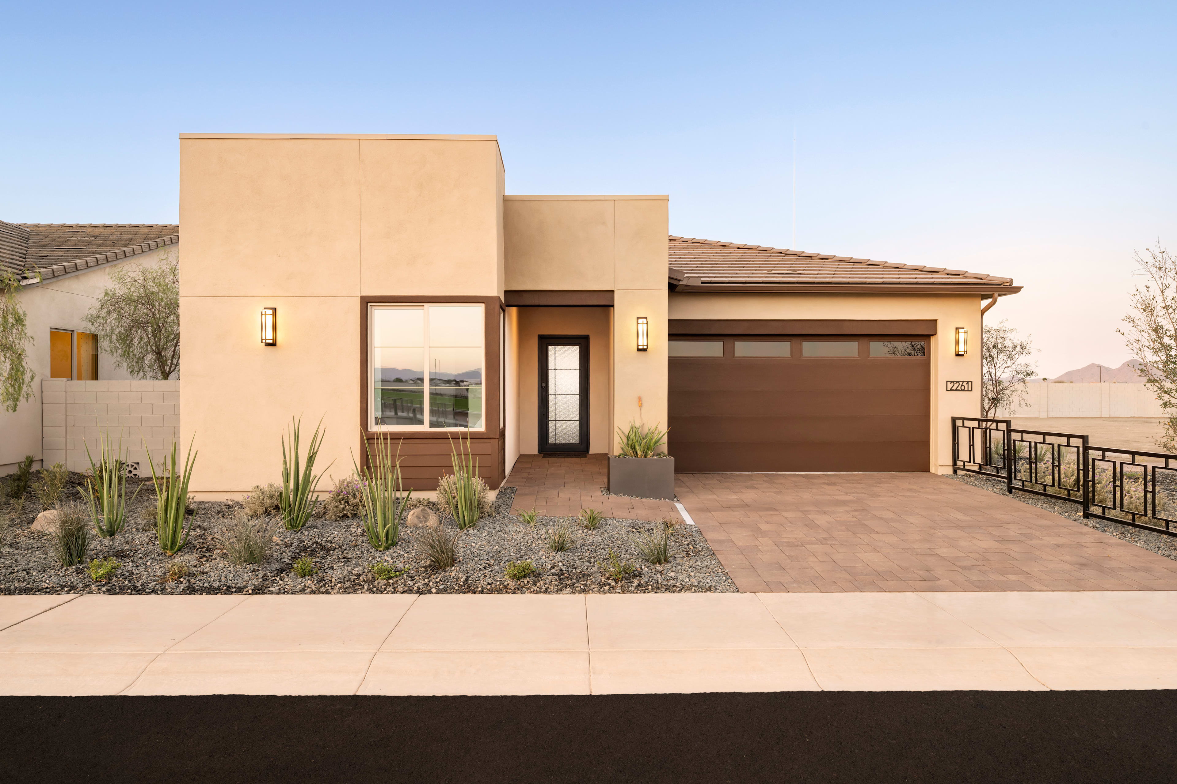 A modern, single-story house with a beige exterior, a garage door, and a landscaped front yard with desert plants against a clear blue sky.