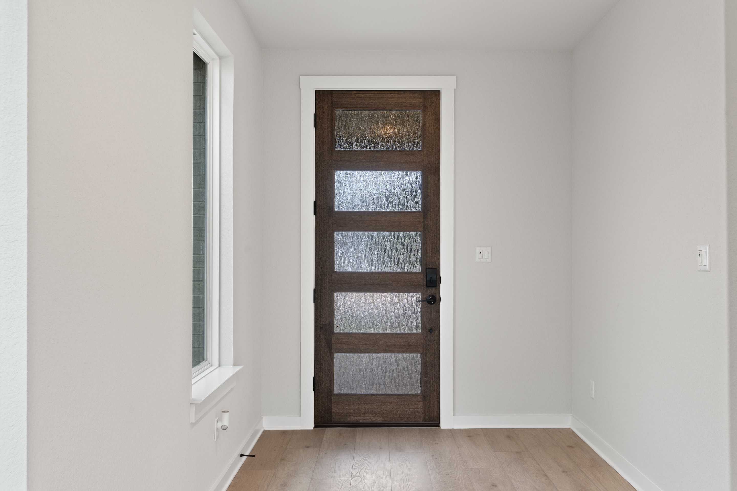 A wooden door with frosted glass panels stands in a bright, minimalist hallway with white walls and light-colored hardwood flooring.