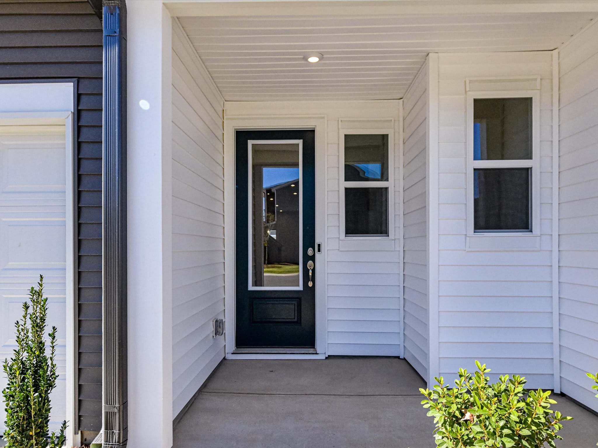 The image shows a white wooden house with a black front door and windows, surrounded by greenery in the foreground.