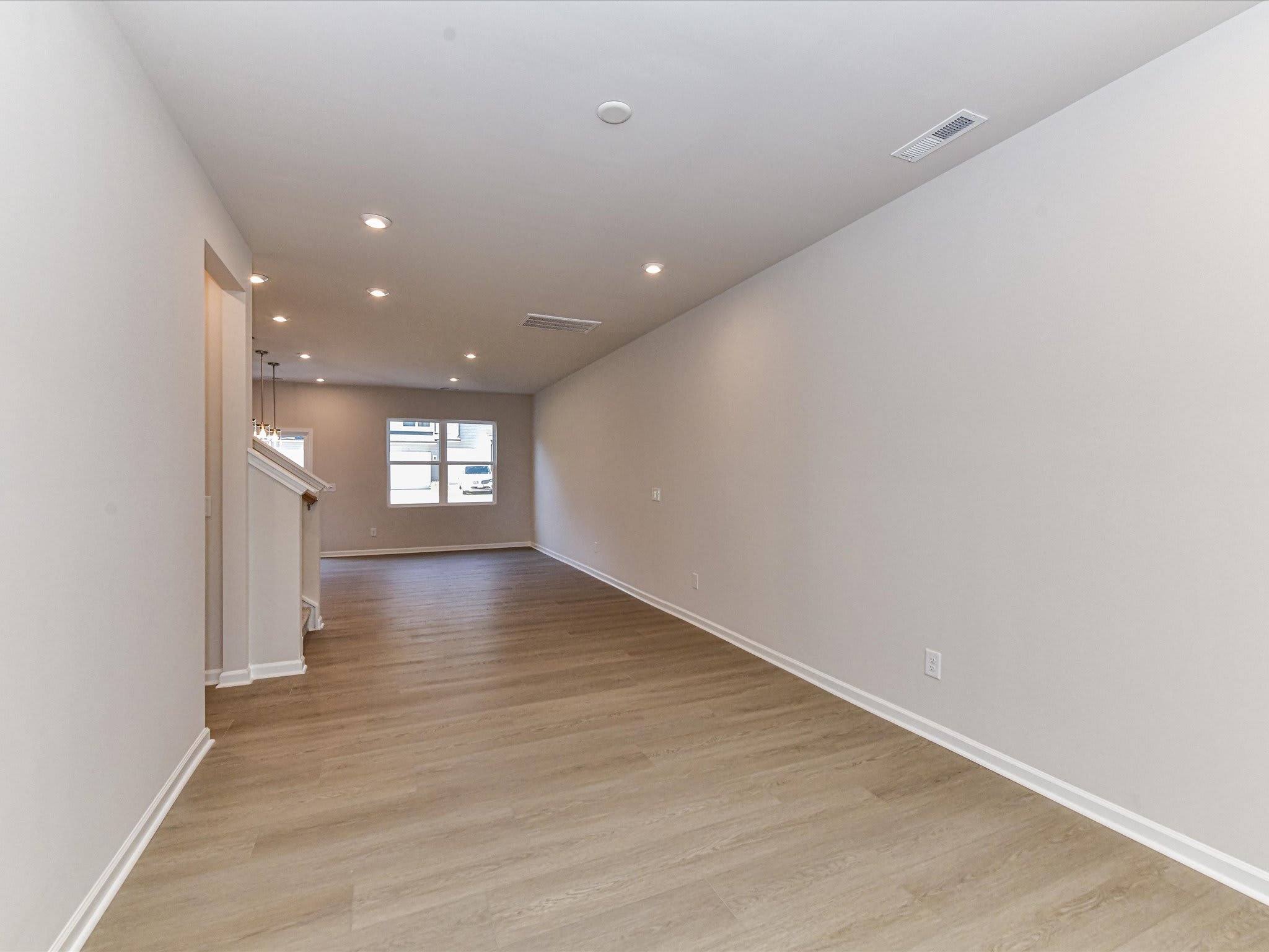 A spacious, well-lit room with a hardwood floor and neutral-colored walls, featuring recessed lighting and a window providing natural illumination.