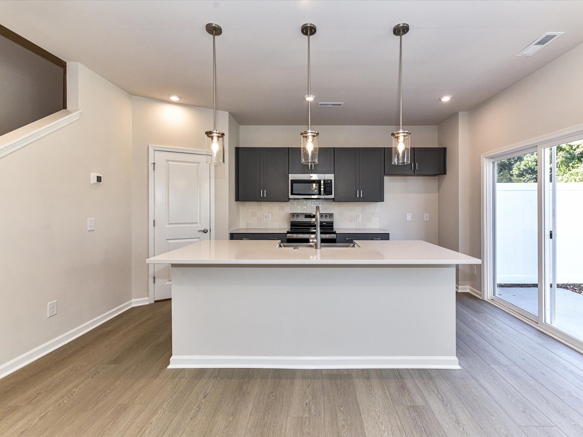 A modern, open-concept kitchen with dark cabinets, a white island, and pendant lighting fixtures, set against a backdrop of hardwood floors and a sliding glass door leading to an outdoor area.