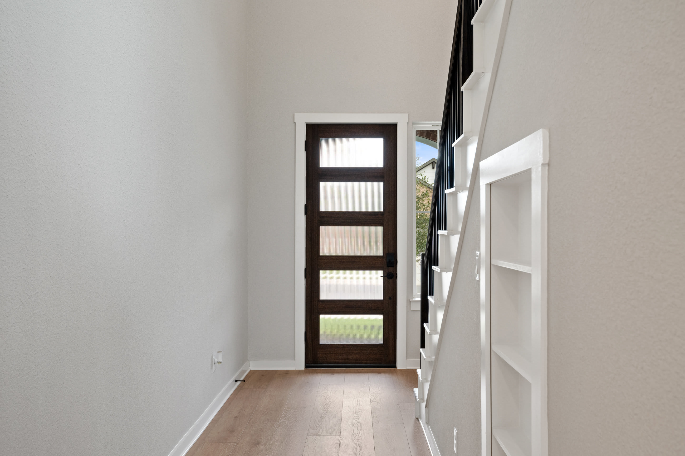 A narrow, dark-framed door with multiple glass panels stands in a bright, white-walled hallway with a wooden floor.