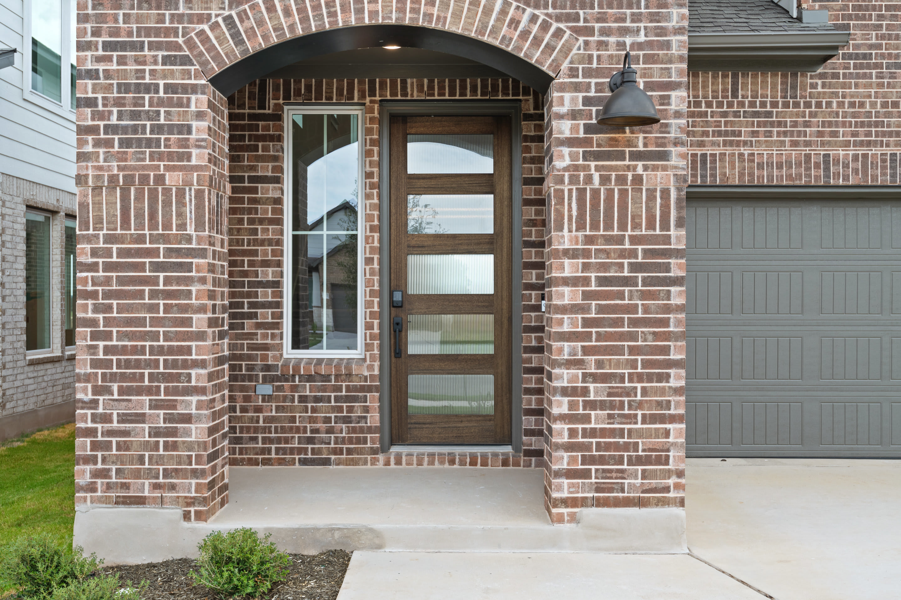 A brick archway frames a wooden front door with glass panels, leading to a paved walkway and a grassy lawn in the background.