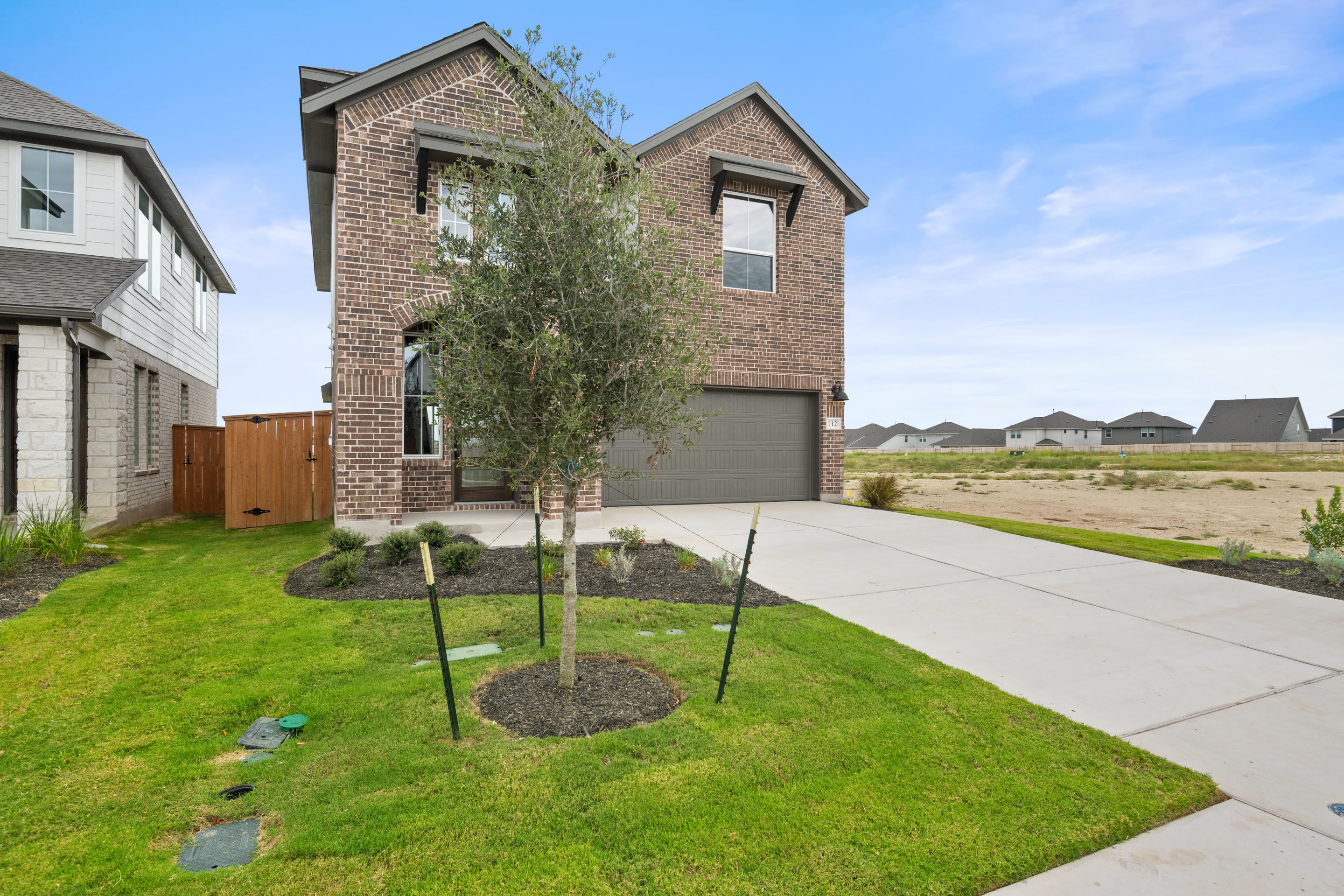 A two-story brick house with a garage, surrounded by a well-manicured lawn and landscaping, stands in a residential neighborhood against a clear blue sky.