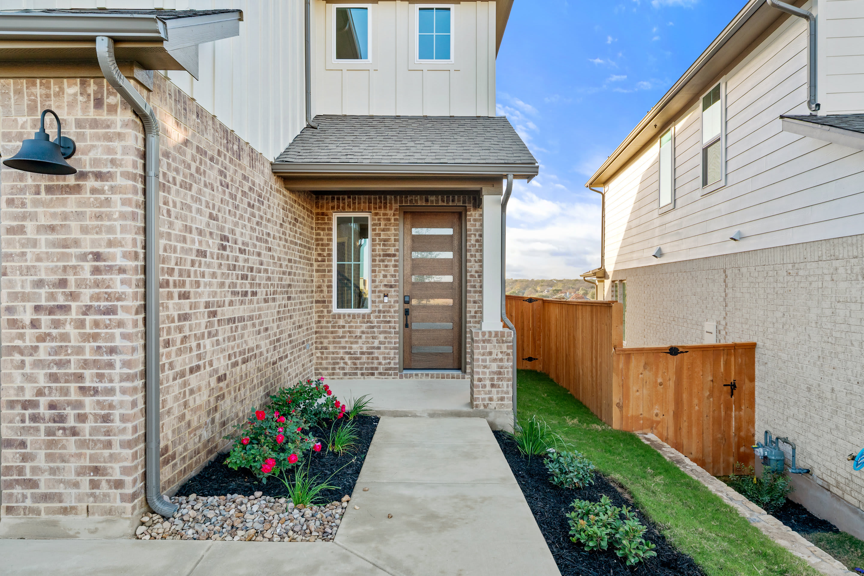A brick and wood-paneled house with a well-maintained front yard, featuring a concrete walkway, flower beds, and a wooden fence in the background.