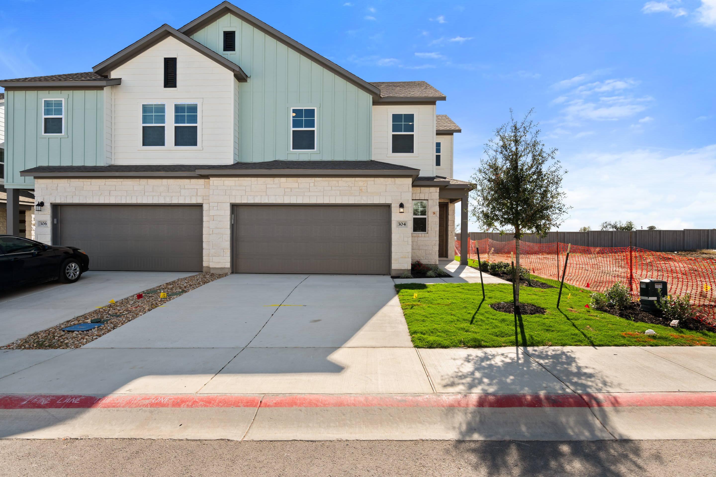 A two-story residential house with a garage, surrounded by a grassy yard and a paved driveway, set against a clear blue sky with some clouds.