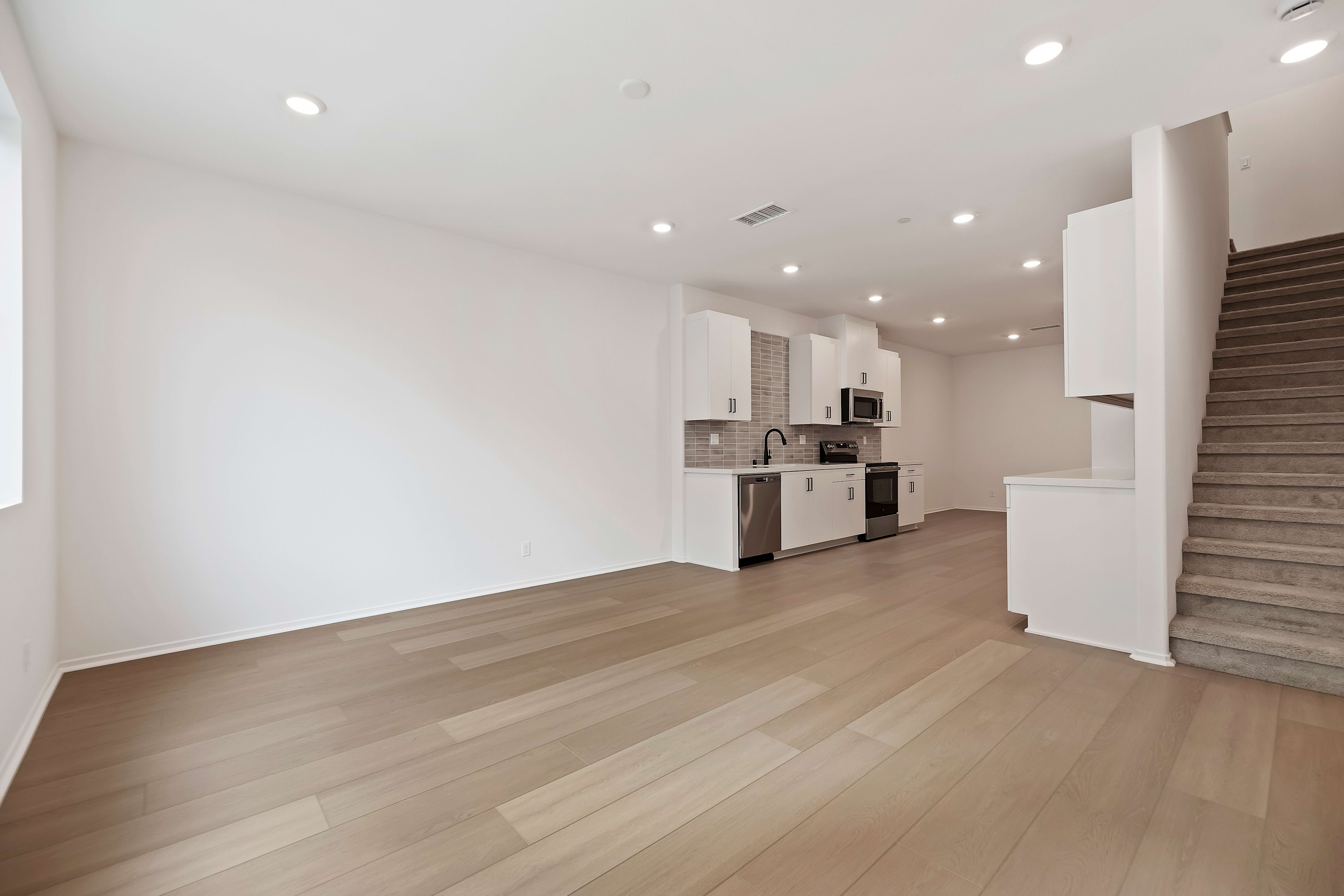 A spacious, open-concept living area with a modern kitchen visible in the background, featuring hardwood floors and recessed lighting throughout.