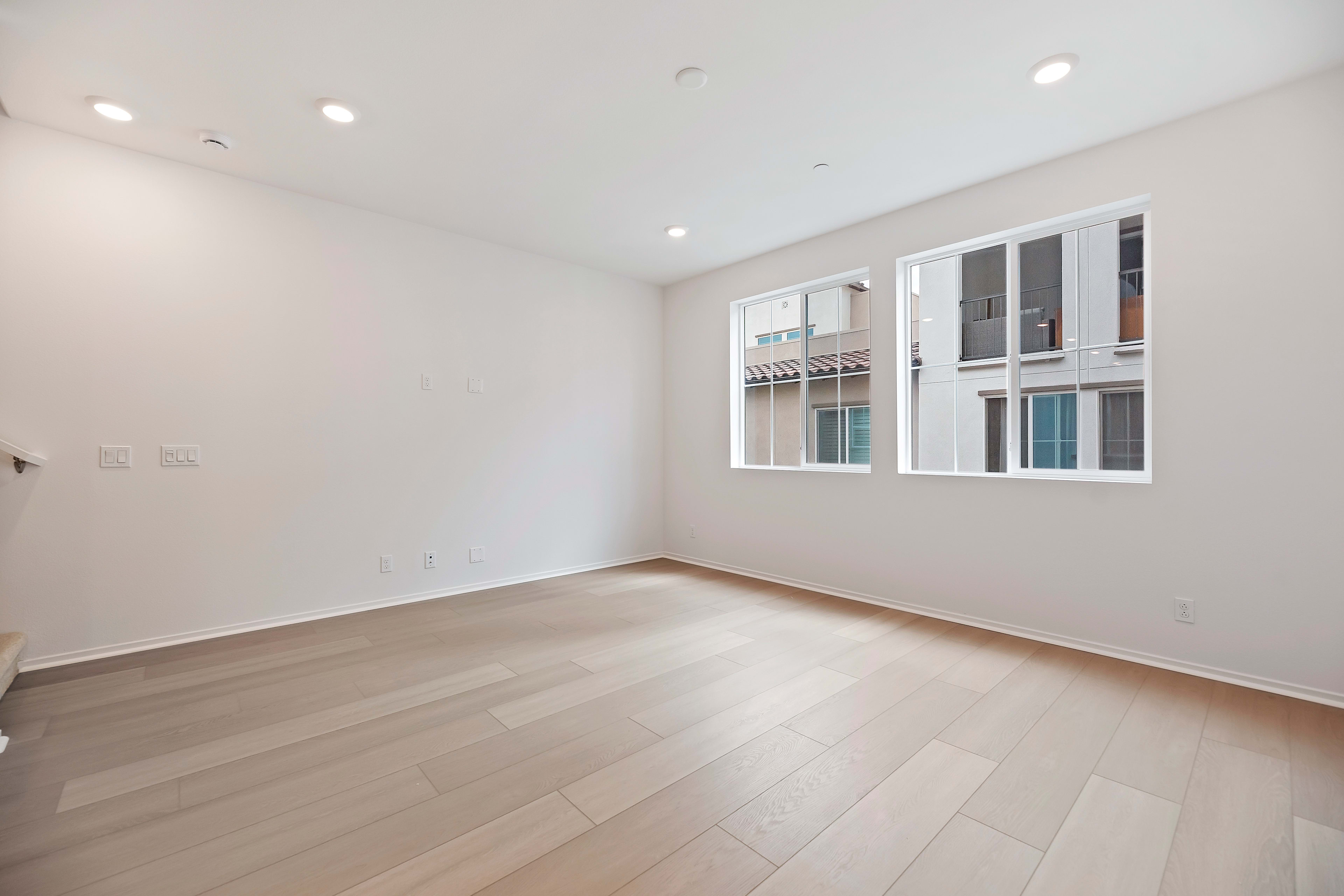 A spacious, well-lit room with white walls, hardwood floors, and large windows overlooking a building exterior.