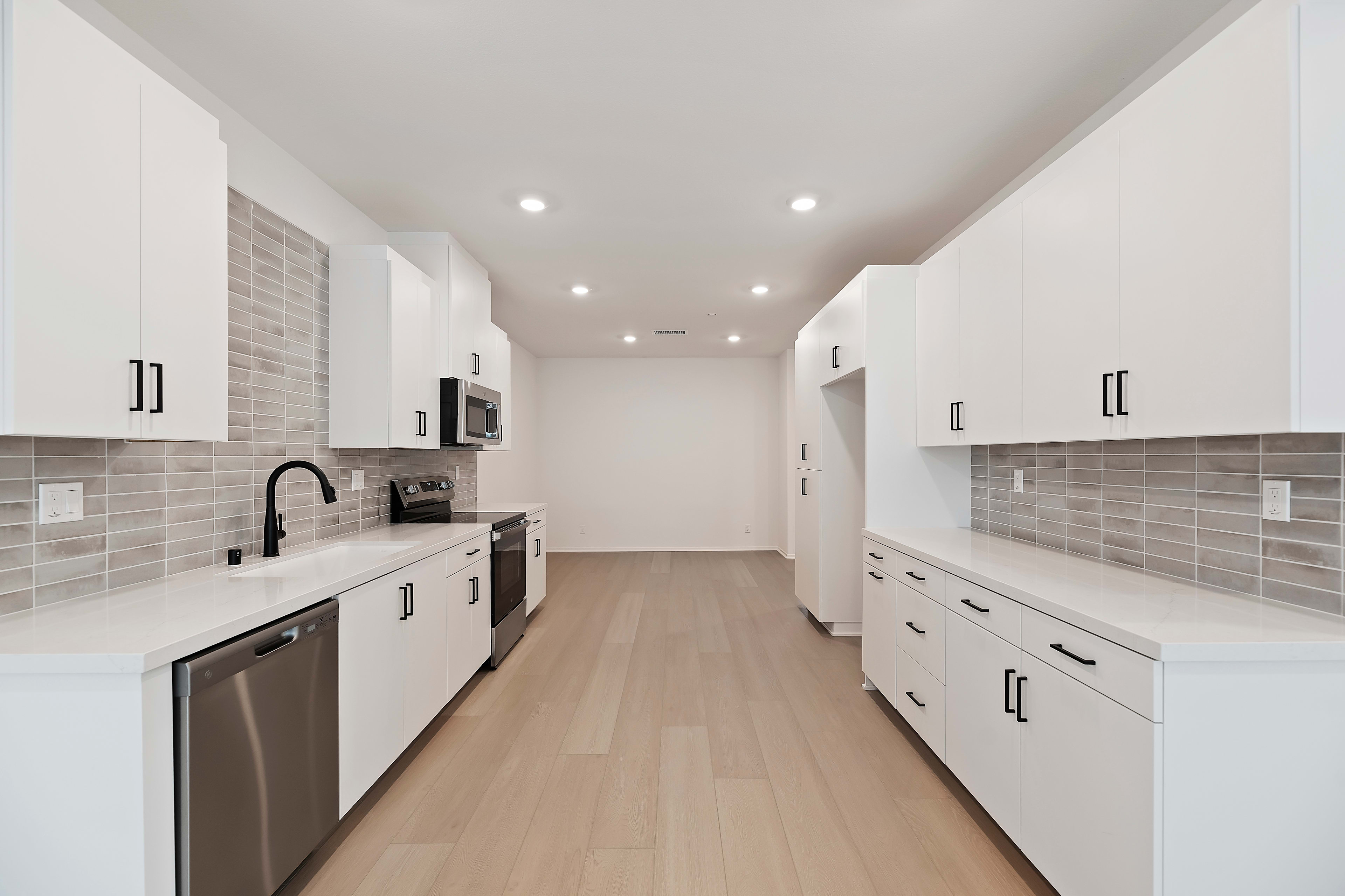A modern, minimalist kitchen with white cabinets, gray tile backsplash, and hardwood floors, creating a clean and spacious atmosphere.