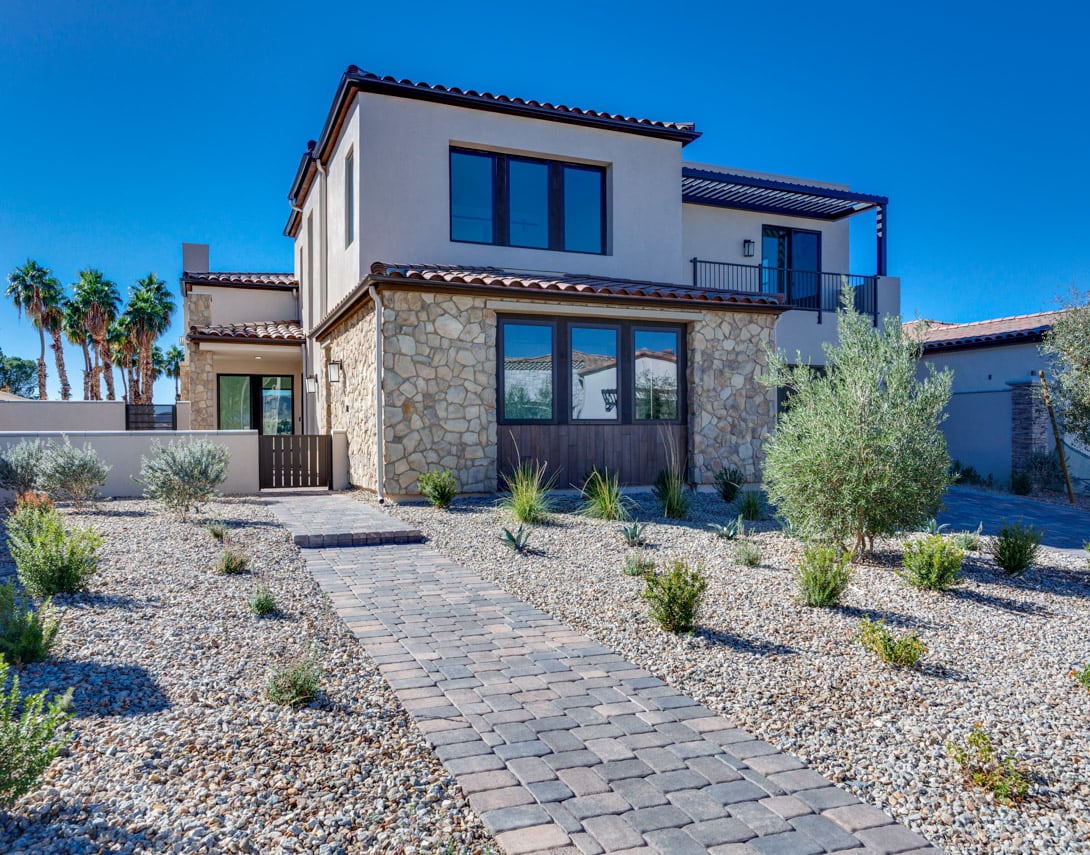 A modern, two-story Mediterranean-style house with a stone exterior, surrounded by a paved walkway and desert landscaping against a clear blue sky.