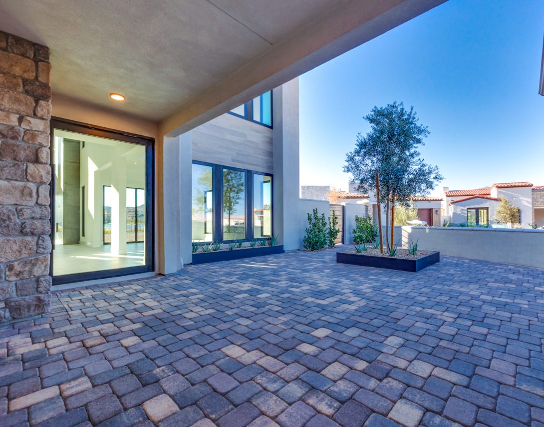 A paved courtyard with brick pavers leads to a modern building with large windows, surrounded by trees and a clear blue sky.