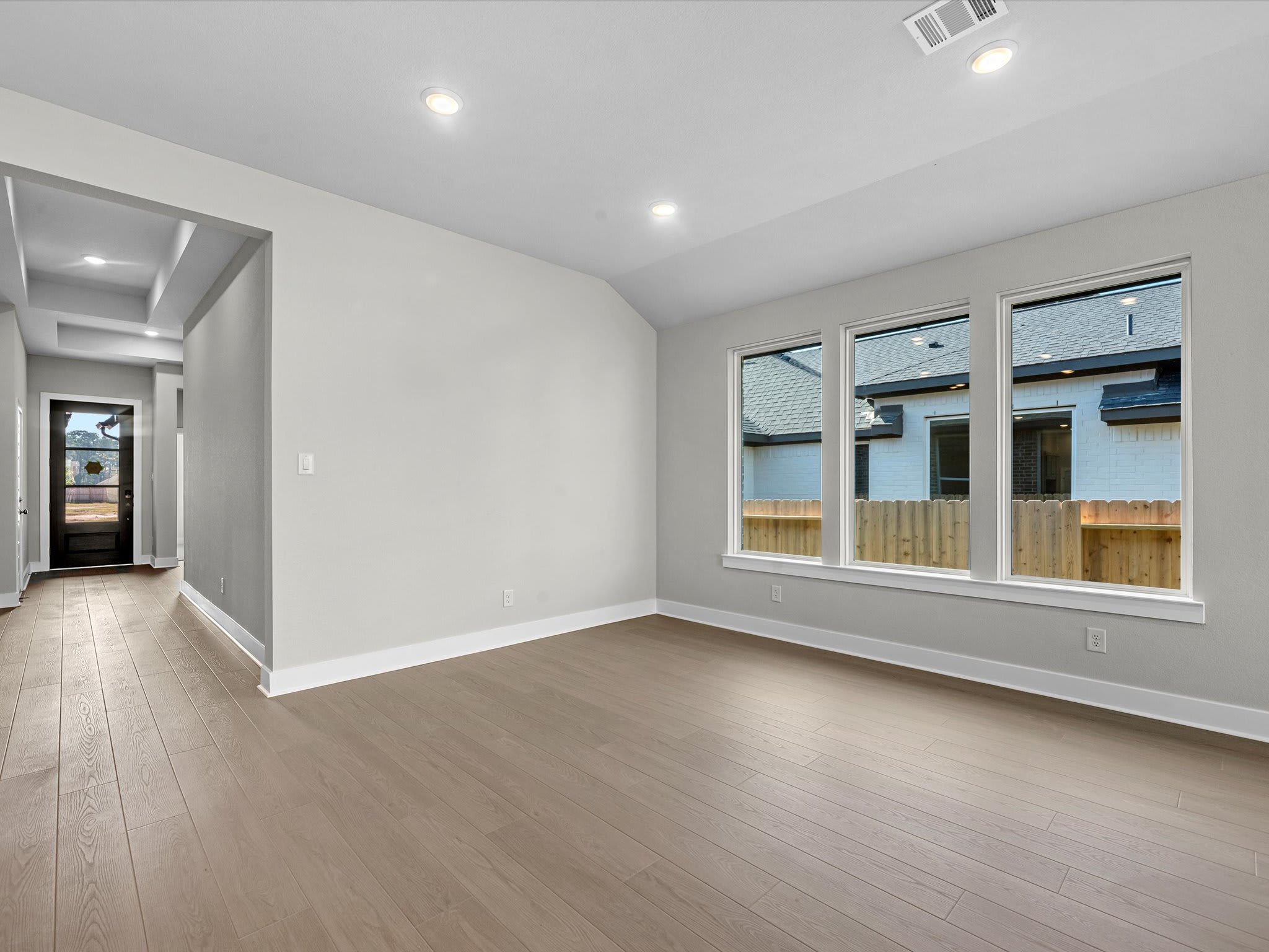 A spacious, well-lit room with a hardwood floor, white walls, and large windows overlooking a fenced outdoor area.