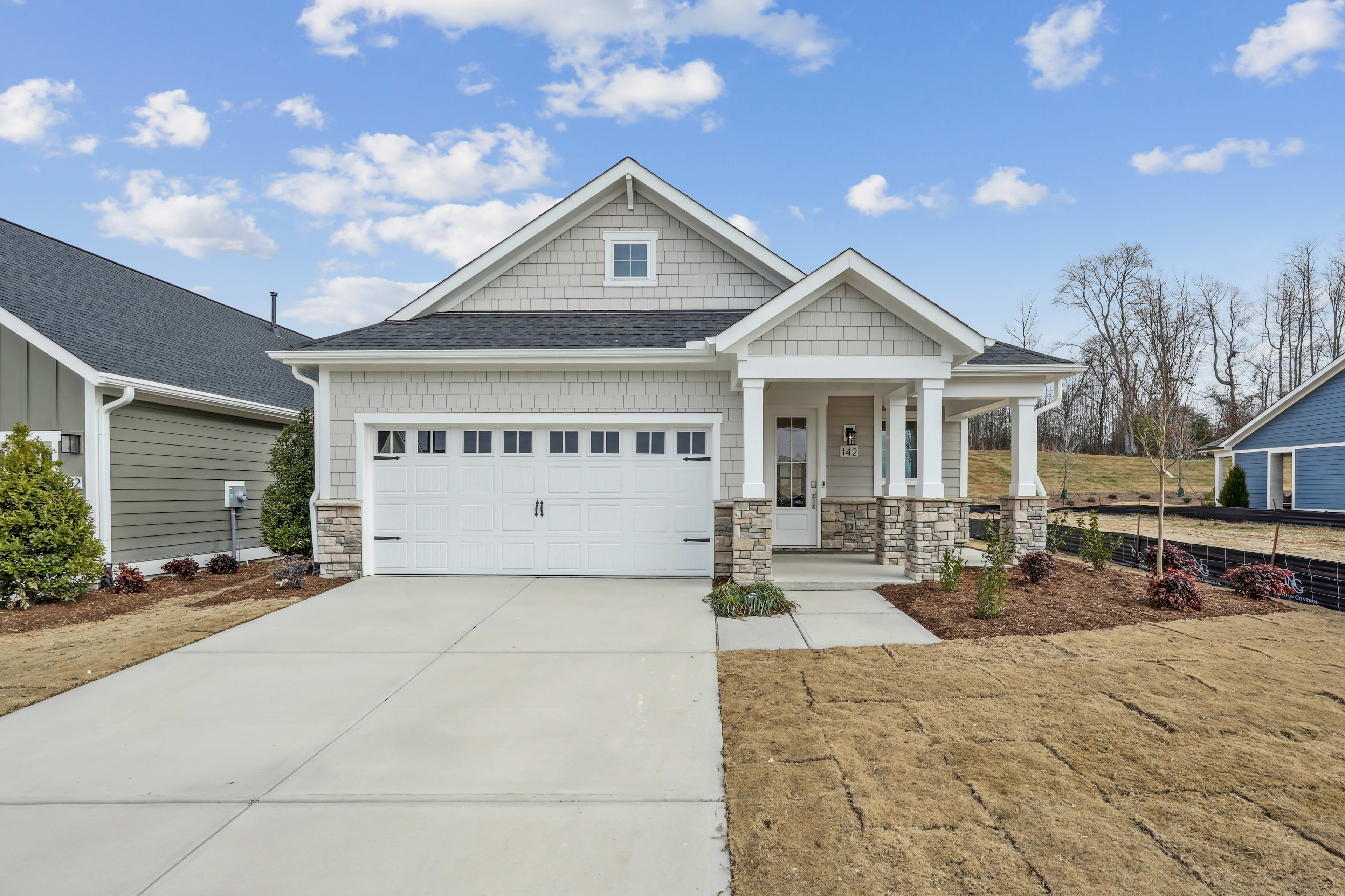 A newly constructed two-story house with a garage, surrounded by a well-manicured lawn and a few trees in the background, set against a blue sky with scattered clouds.