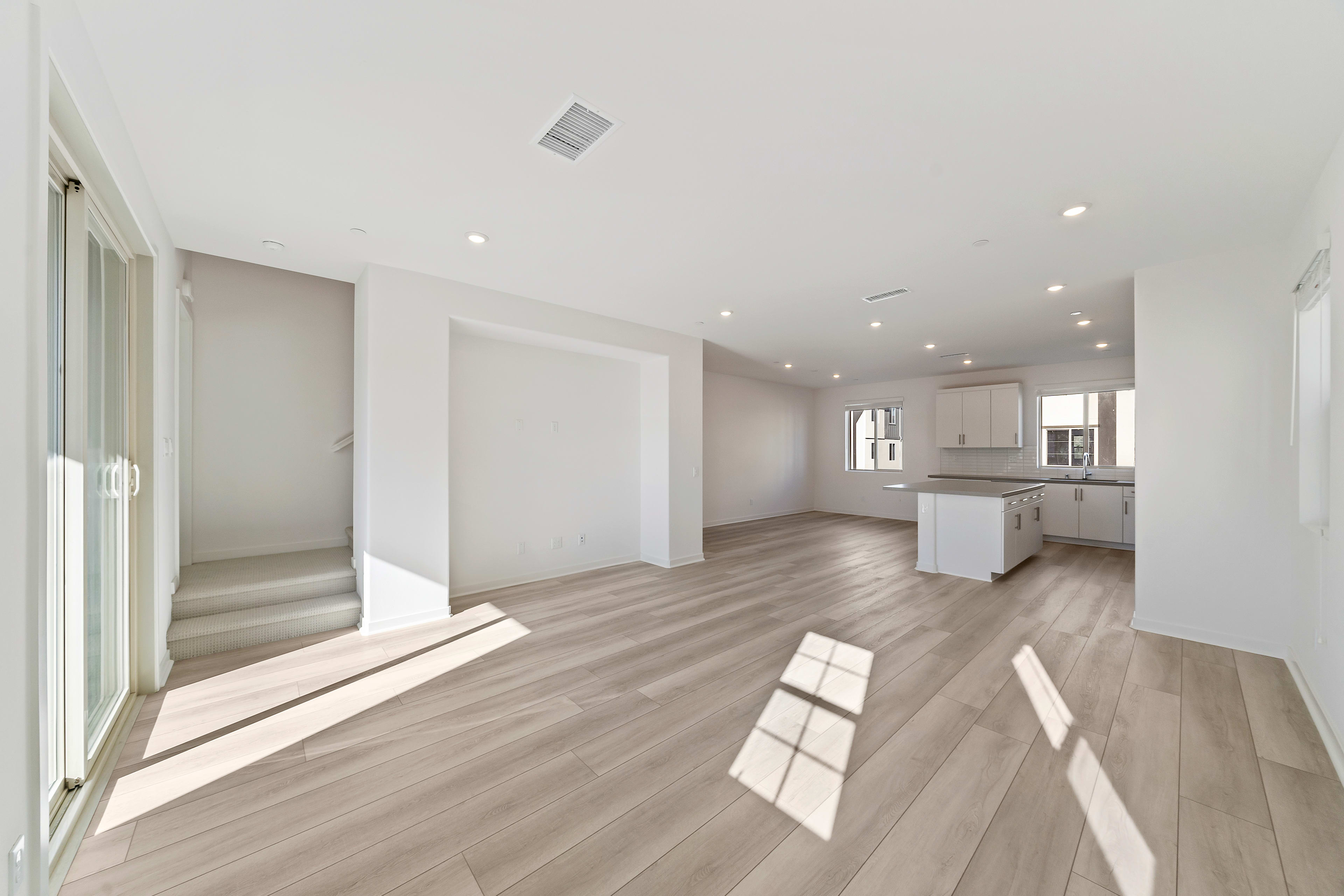 A spacious, modern interior with light-colored hardwood floors, white walls, and recessed lighting, featuring an open kitchen area with white cabinets and countertops.