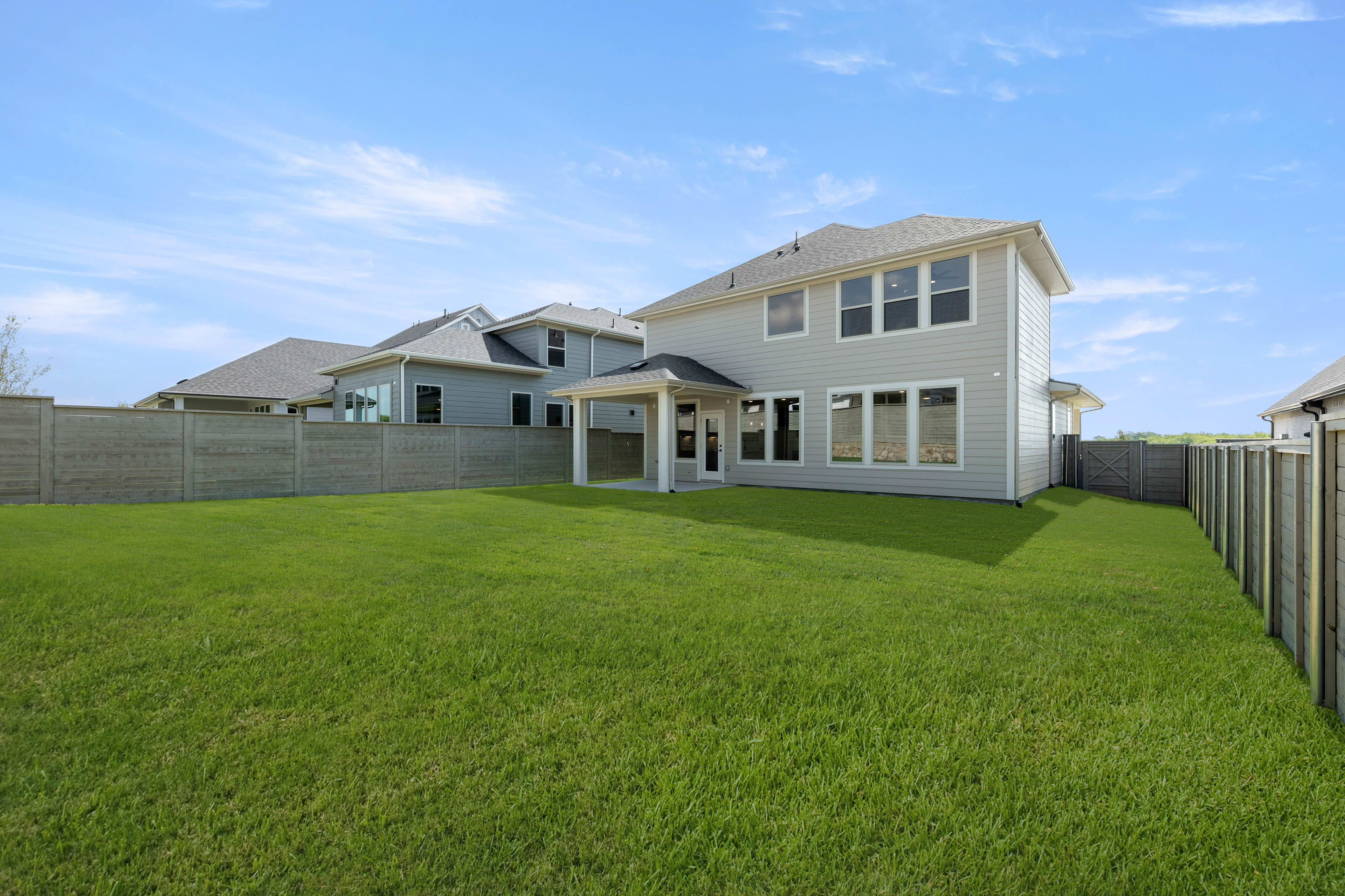 A well-manicured lawn stretches out in the foreground, leading up to a two-story residential home with a porch and multiple windows, set against a clear blue sky with wispy clouds.