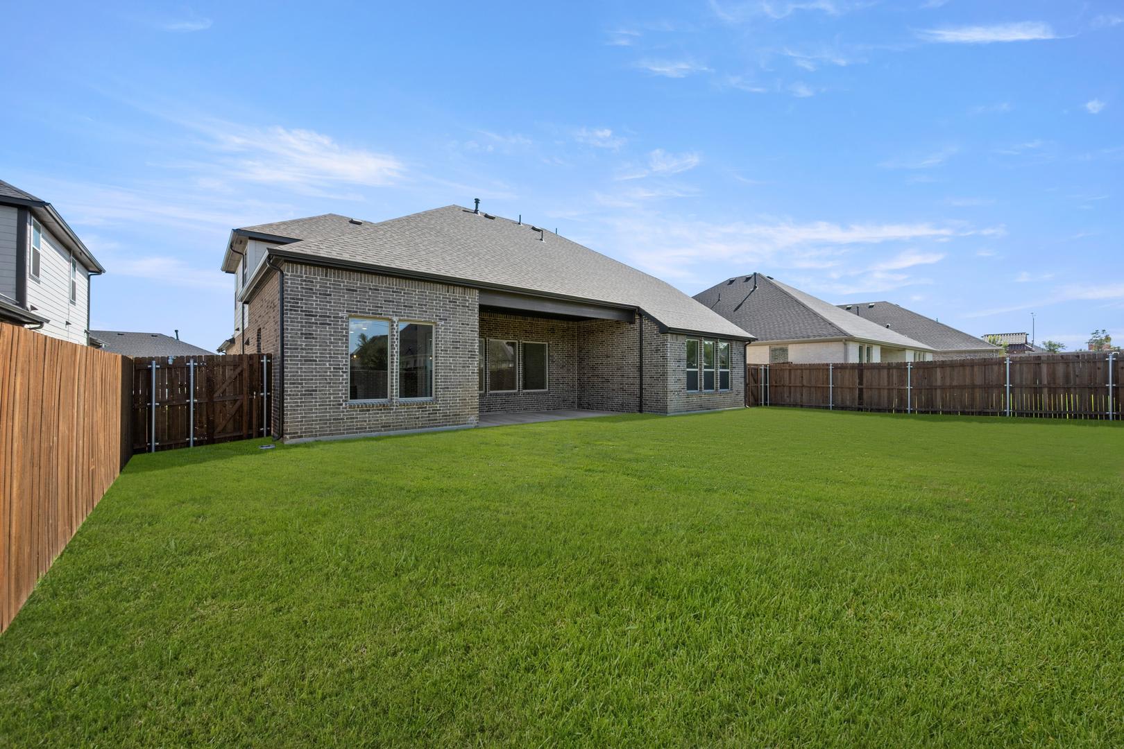 A well-manicured lawn stretches out in the foreground, leading up to a brick and siding residential home with a slanted roof in the background, set against a clear blue sky.