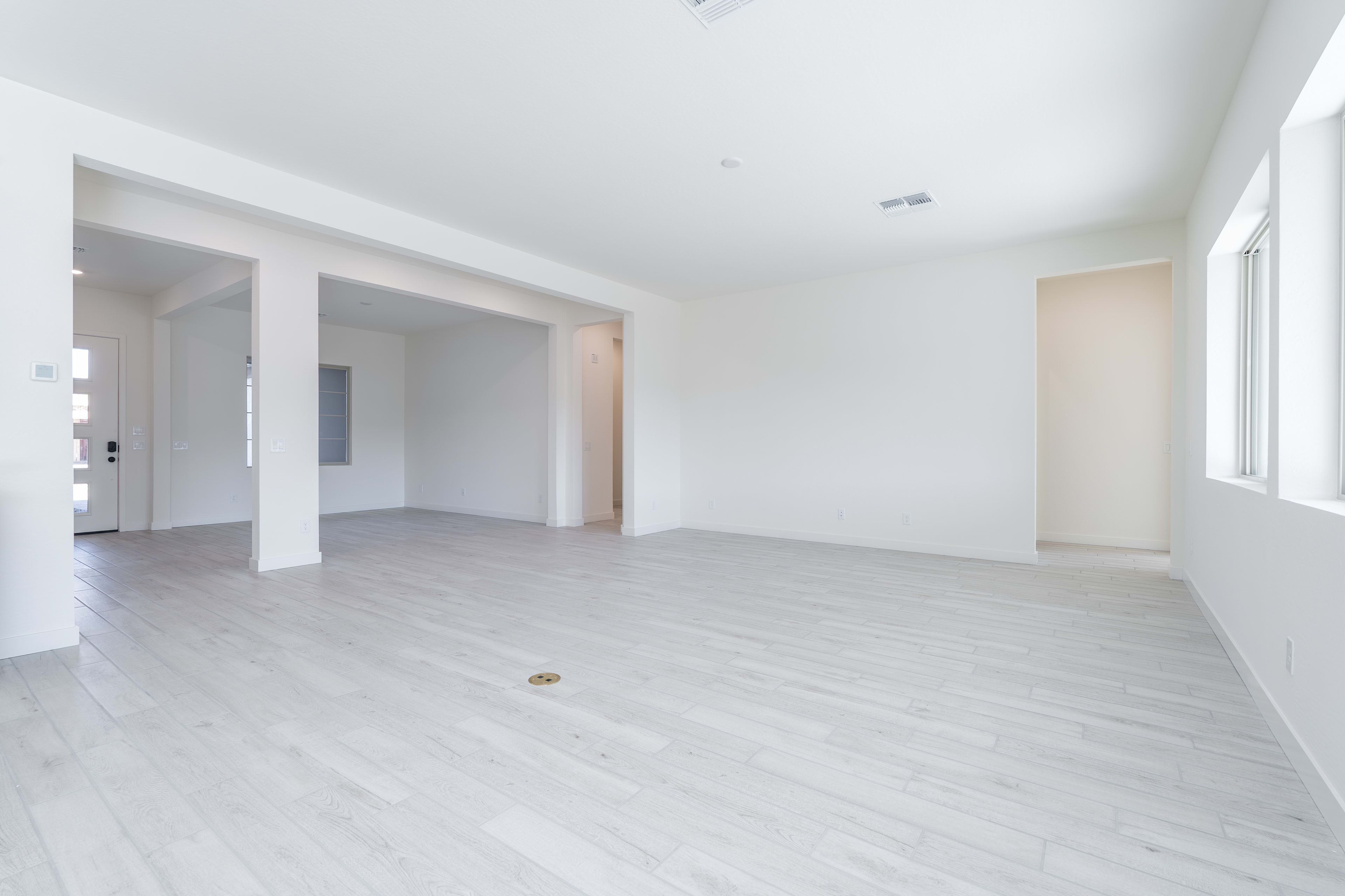 A spacious, minimalist room with white walls, light-colored hardwood floors, and open doorways leading to other areas of the space.