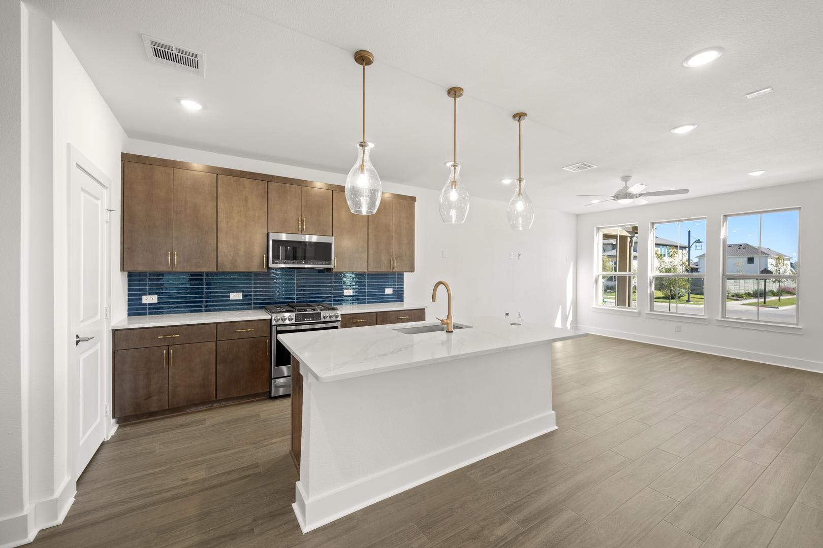 A modern and spacious kitchen with wooden cabinets, a white island, and pendant lights, set against a bright and airy living space with large windows.