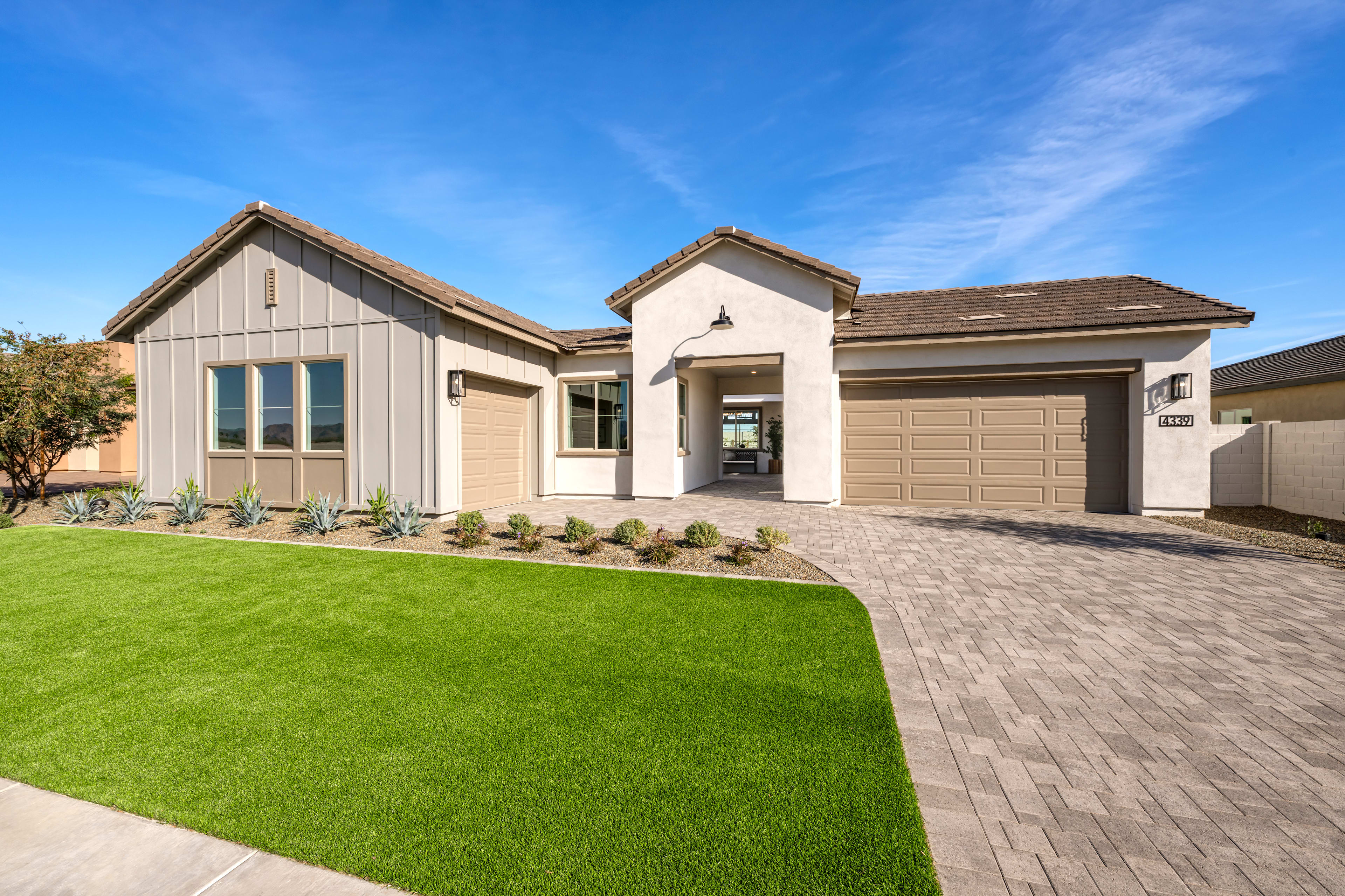 A well-manicured lawn leads to a modern, two-story house with a garage, featuring a mix of siding materials and a tiled roof against a clear blue sky.
