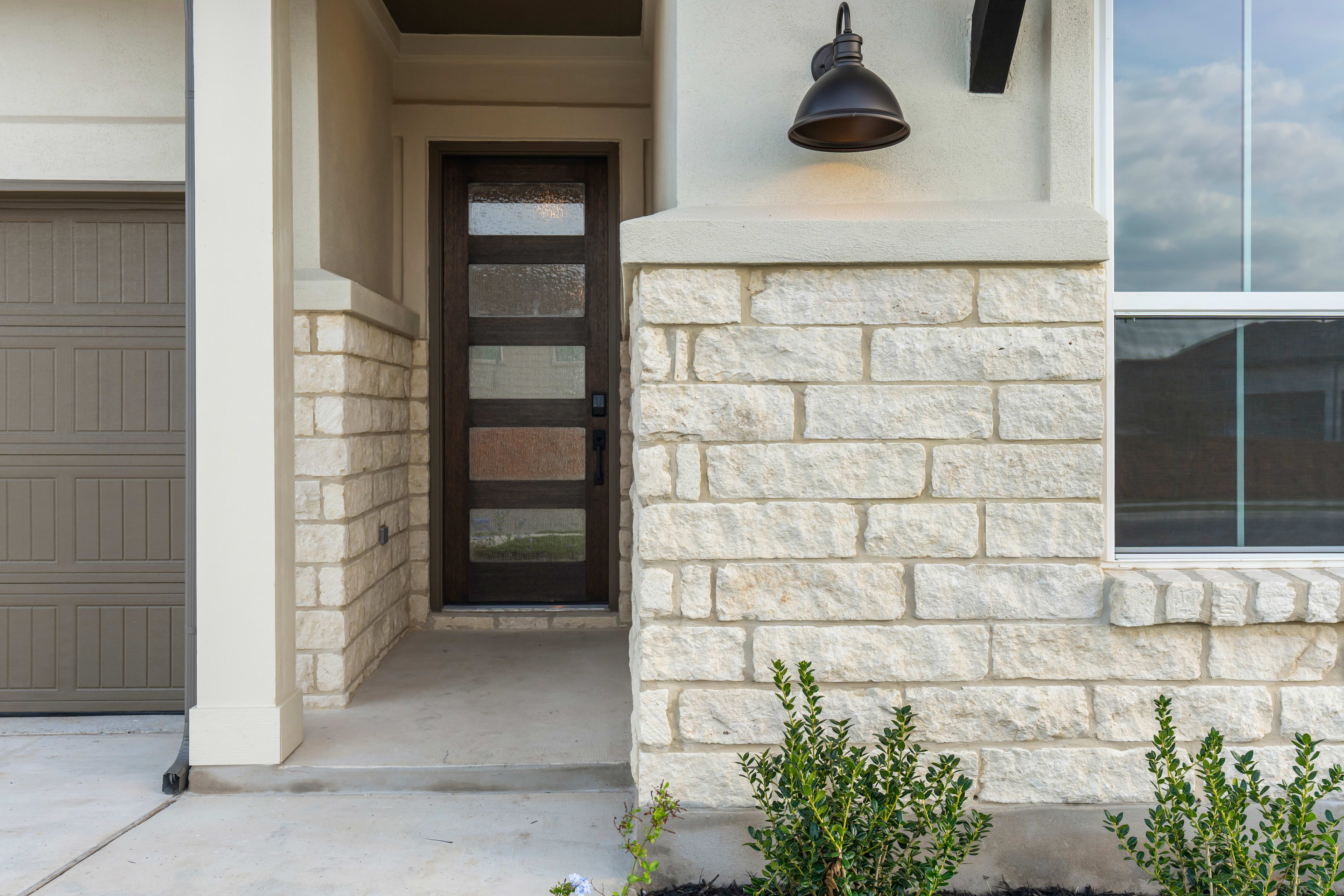 A stone-clad entryway with a black light fixture and a small plant growing near the doorway, set against a cloudy sky background.