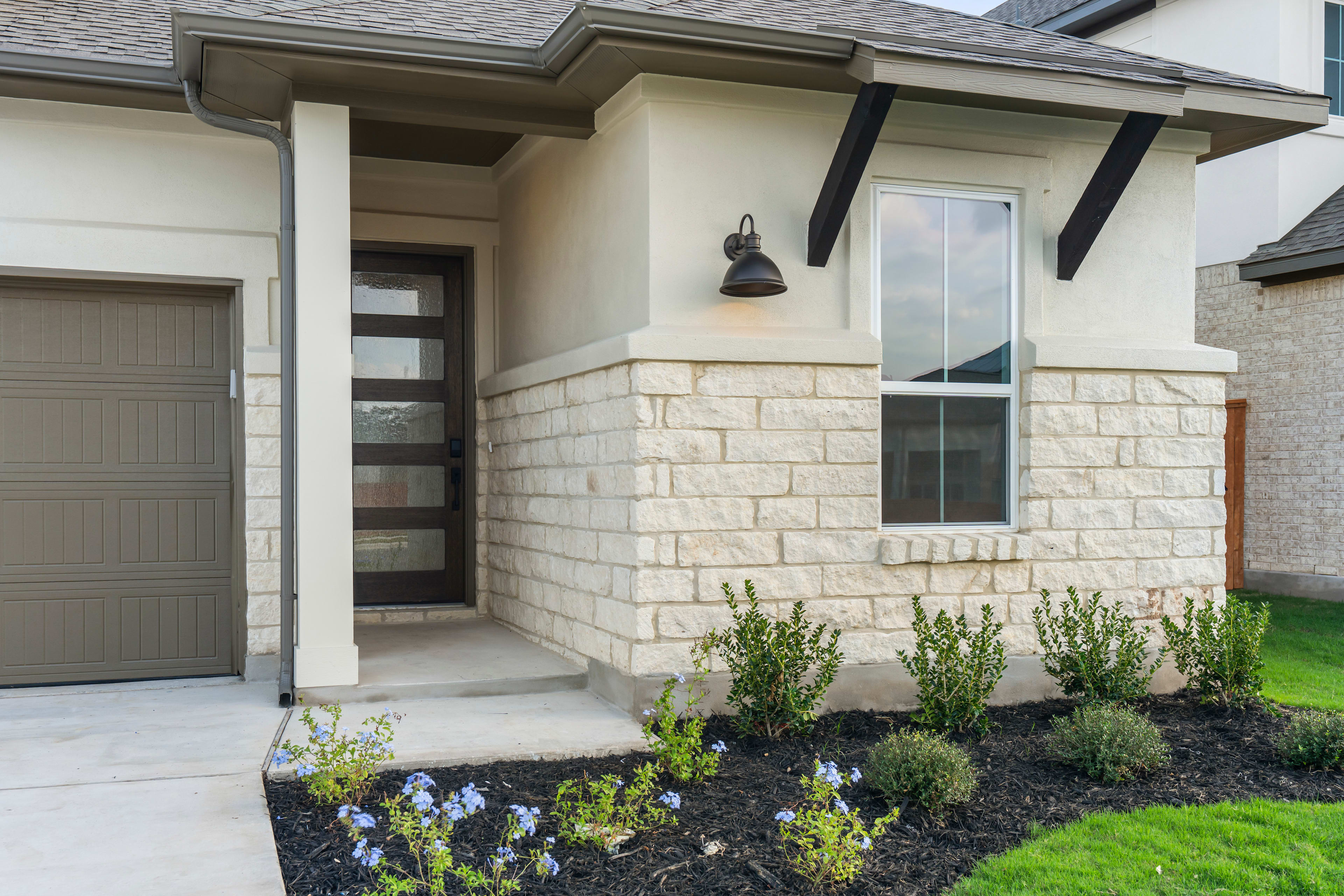 The image shows the front entrance of a modern, two-story house with a stone facade, a covered porch, and a well-maintained landscaped area in the foreground featuring various flowering plants and shrubs.