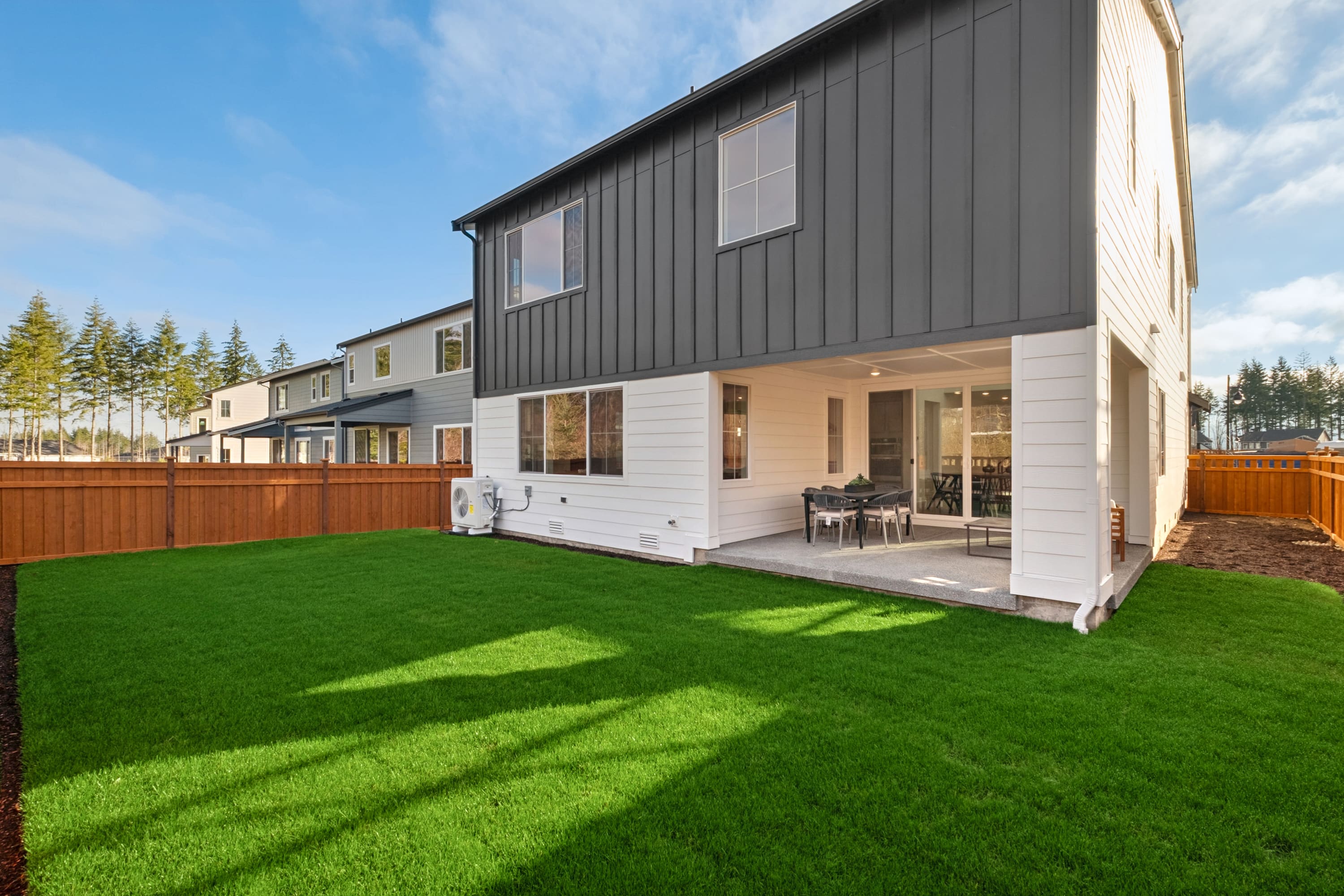 A modern two-story house with a gray exterior and a large backyard covered in lush green grass, surrounded by pine trees in the background.