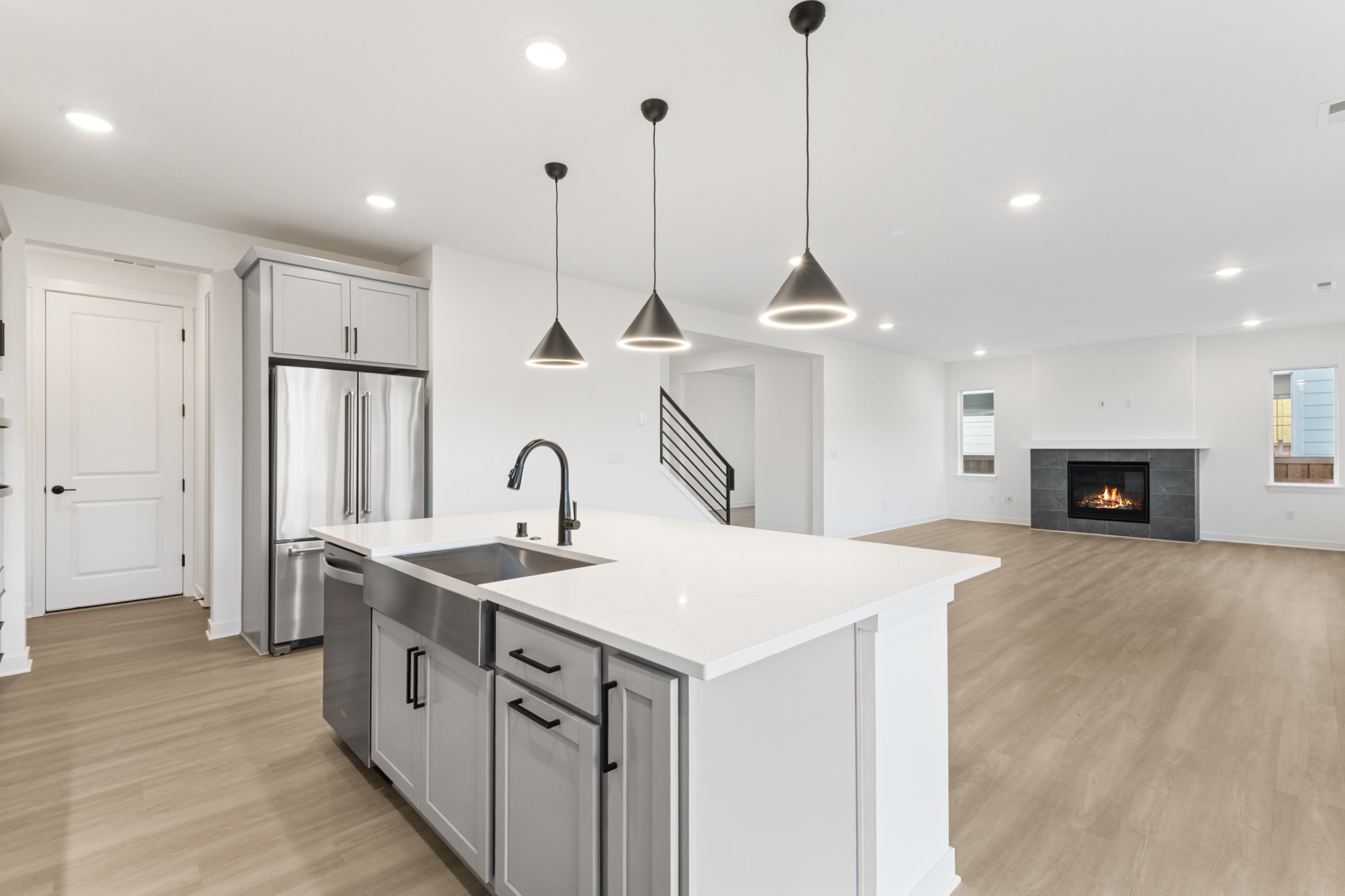 A modern, open-concept kitchen with a white island, stainless steel appliances, and pendant lighting fixtures, set against a backdrop of a living room with a fireplace.